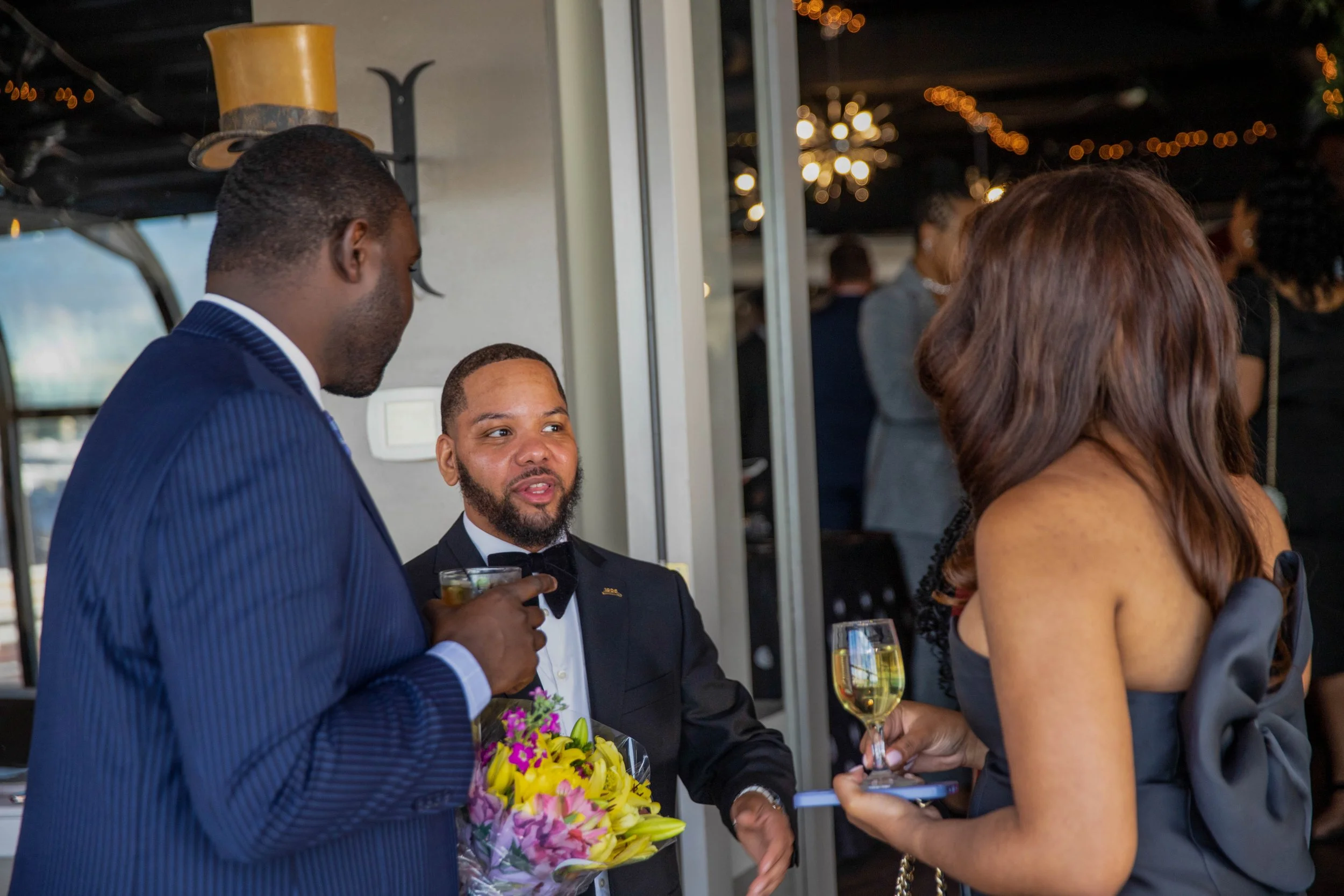People dressed in formal attire at a social event, with one person holding a bouquet of flowers and another holding a glass of wine, indoors with warm lighting and decorations.