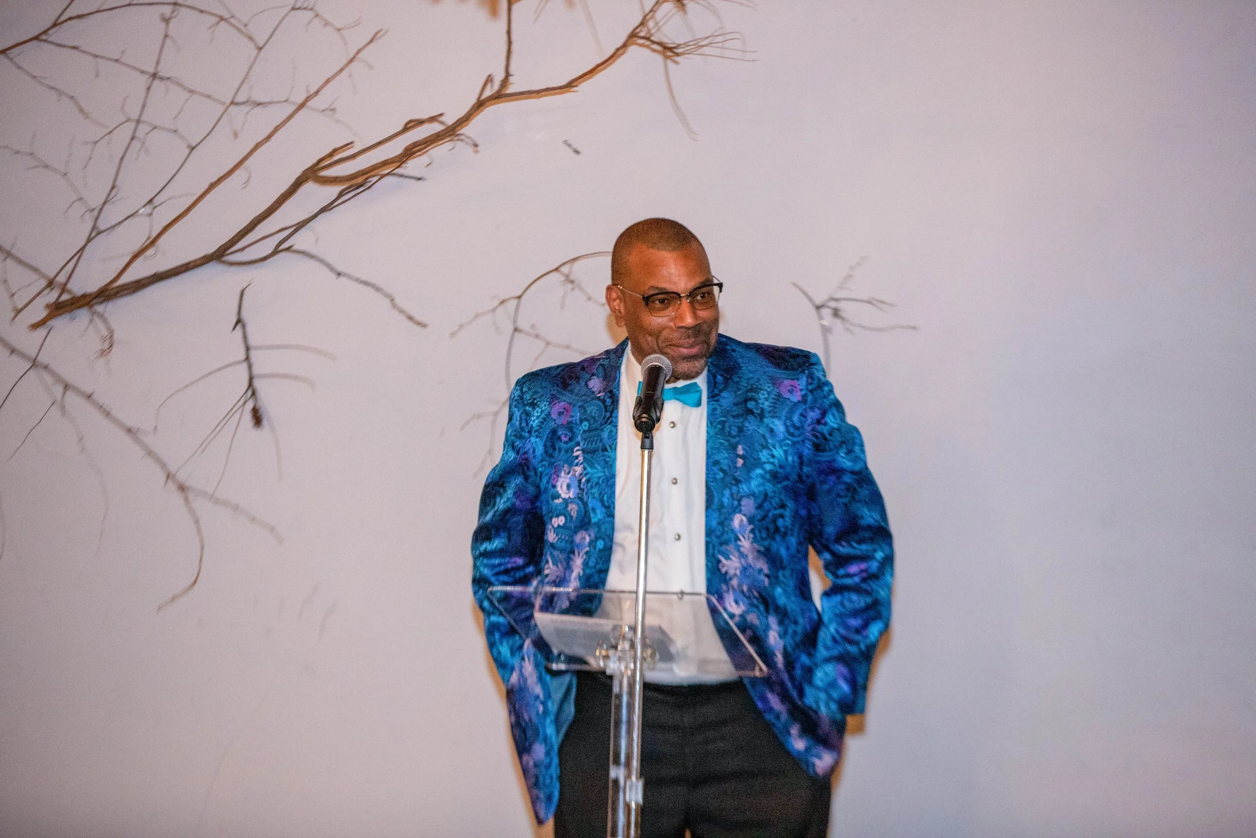 A man in a colorful blue and purple patterned jacket, white shirt, and black pants standing behind a clear podium, speaking into a microphone, with a plain wall and dried branches in the background.