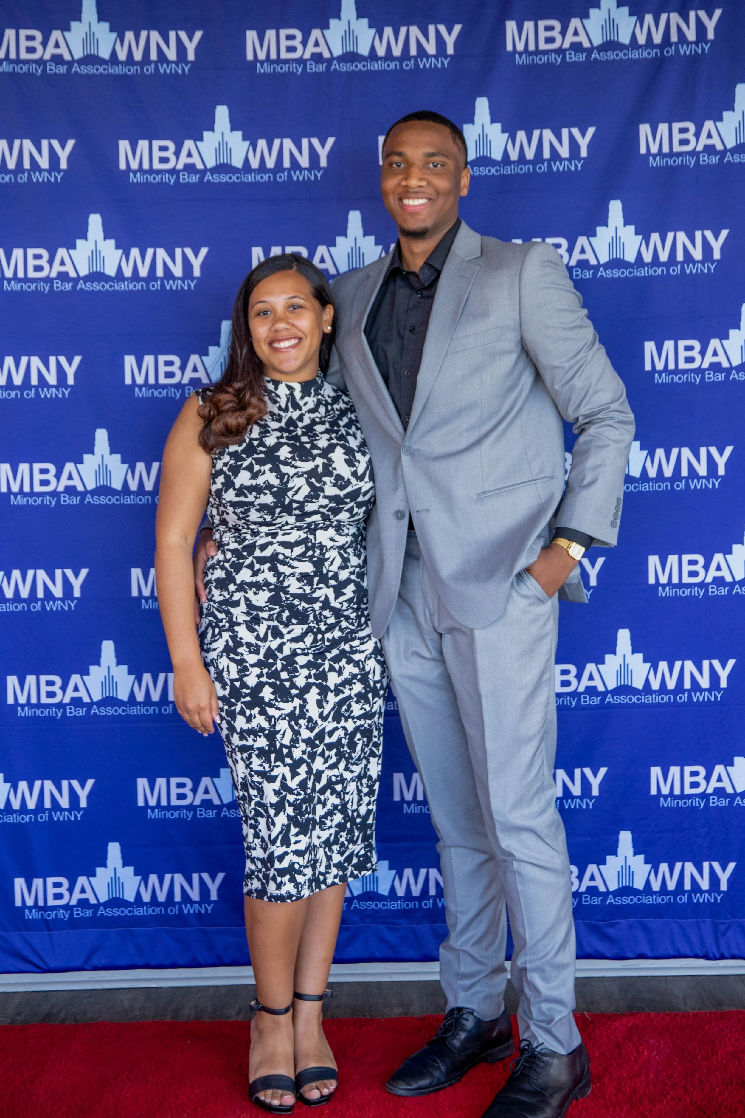 A man and a woman standing together on a red carpet in front of a blue backdrop with the logo and name of the Minority Bar Association of WNY. They are dressed in formal attire, smiling for the photo.
