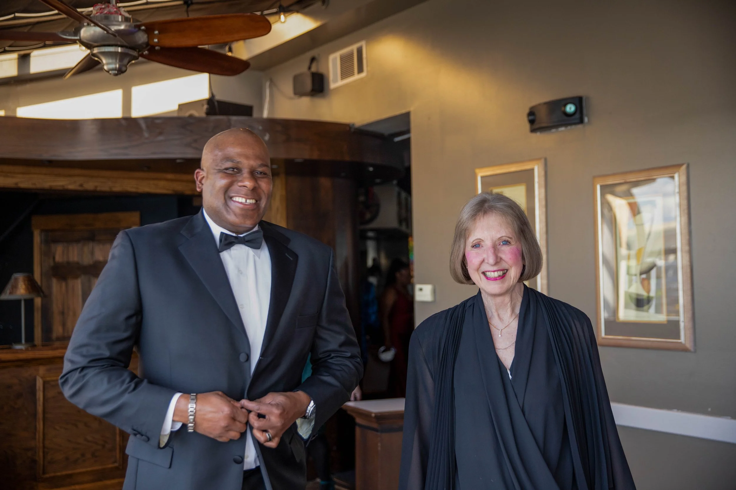 A man in a tuxedo shaking hands with a woman in a black dress in a warmly lit restaurant or event space.