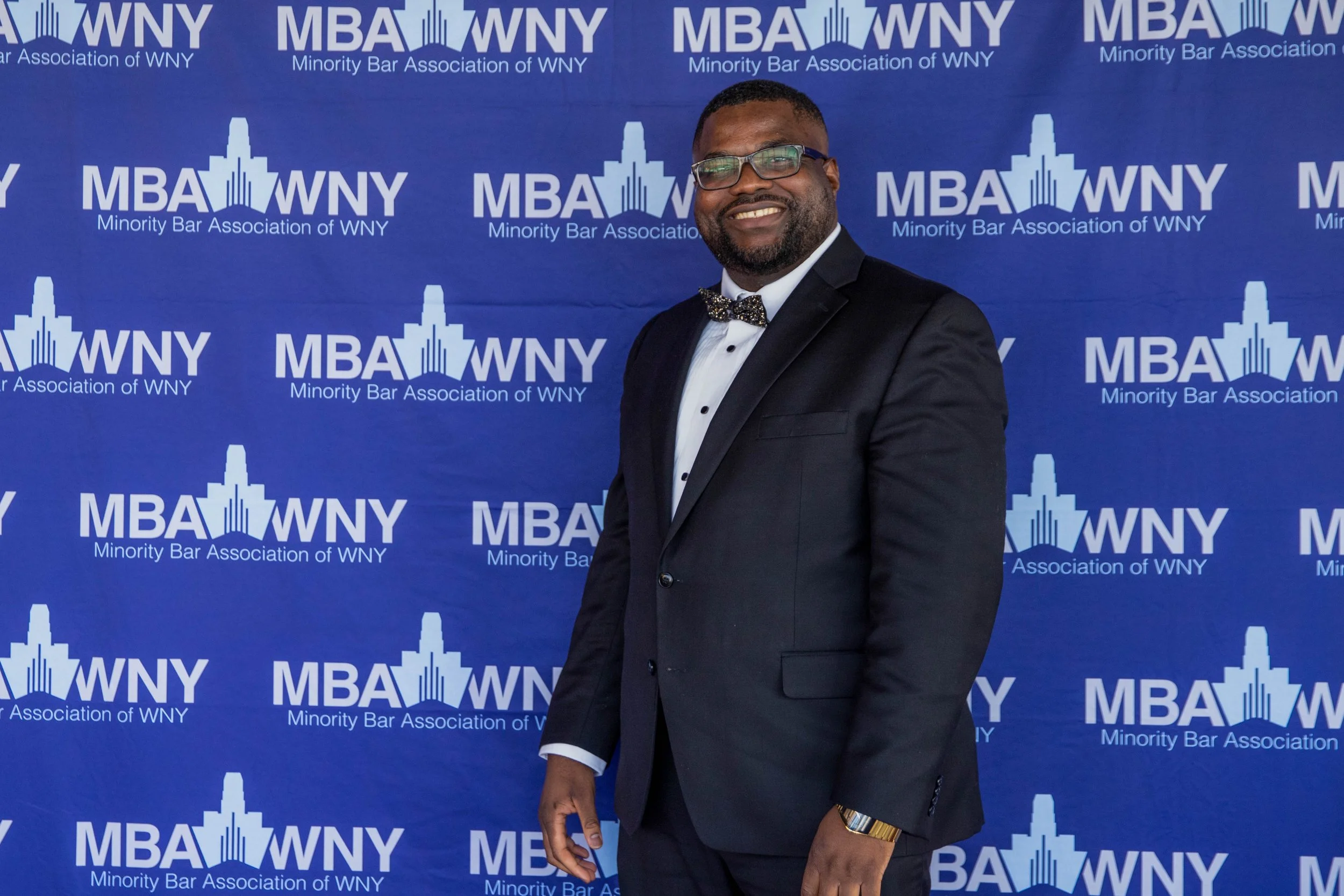 A man in a tuxedo smiling at a formal event, standing in front of a blue backdrop with the logo for the Minority Bar Association of WNY.