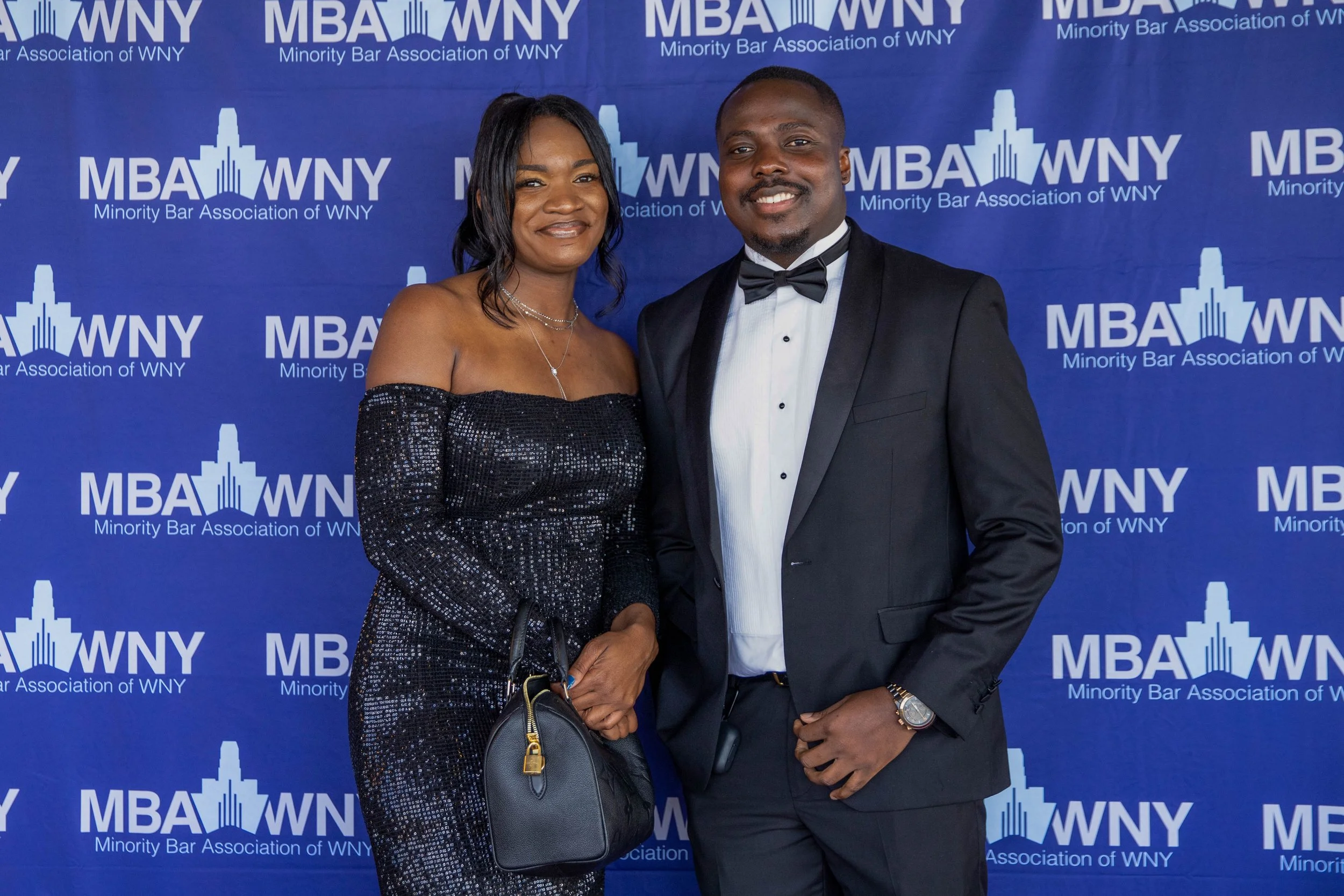 A man and woman in formal attire standing in front of a blue backdrop with the logo and text of the Minority Bar Association of WNY. The woman is wearing a black, off-the-shoulder, sparkly dress and the man is wearing a black tuxedo with a white shir