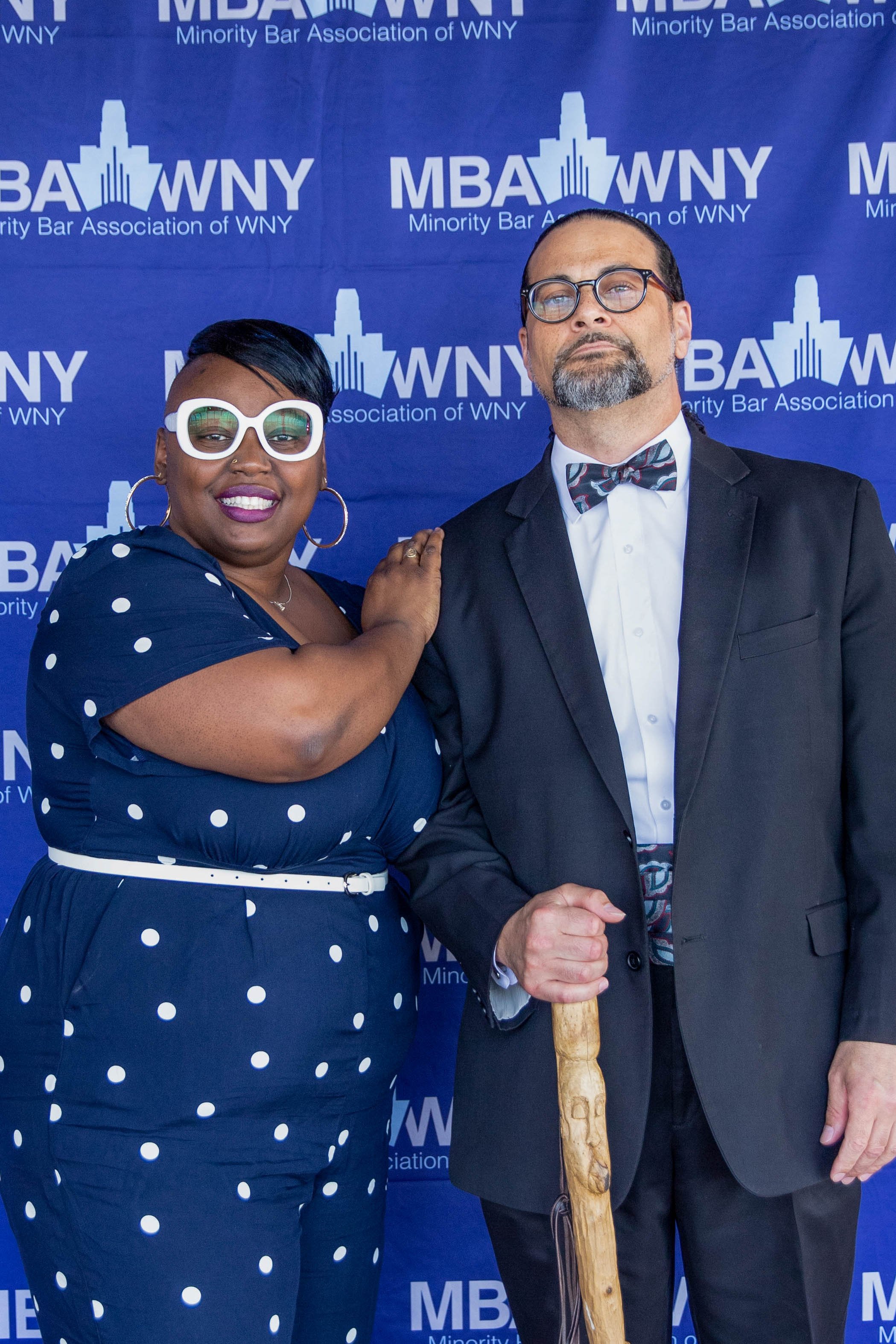 A woman with short black hair, large white sunglasses, and purple lipstick, standing next to a man with glasses, a beard, and a black suit, holding a wooden cane, in front of a blue banner with white text that reads 'Minority Bar Association of WNY' 