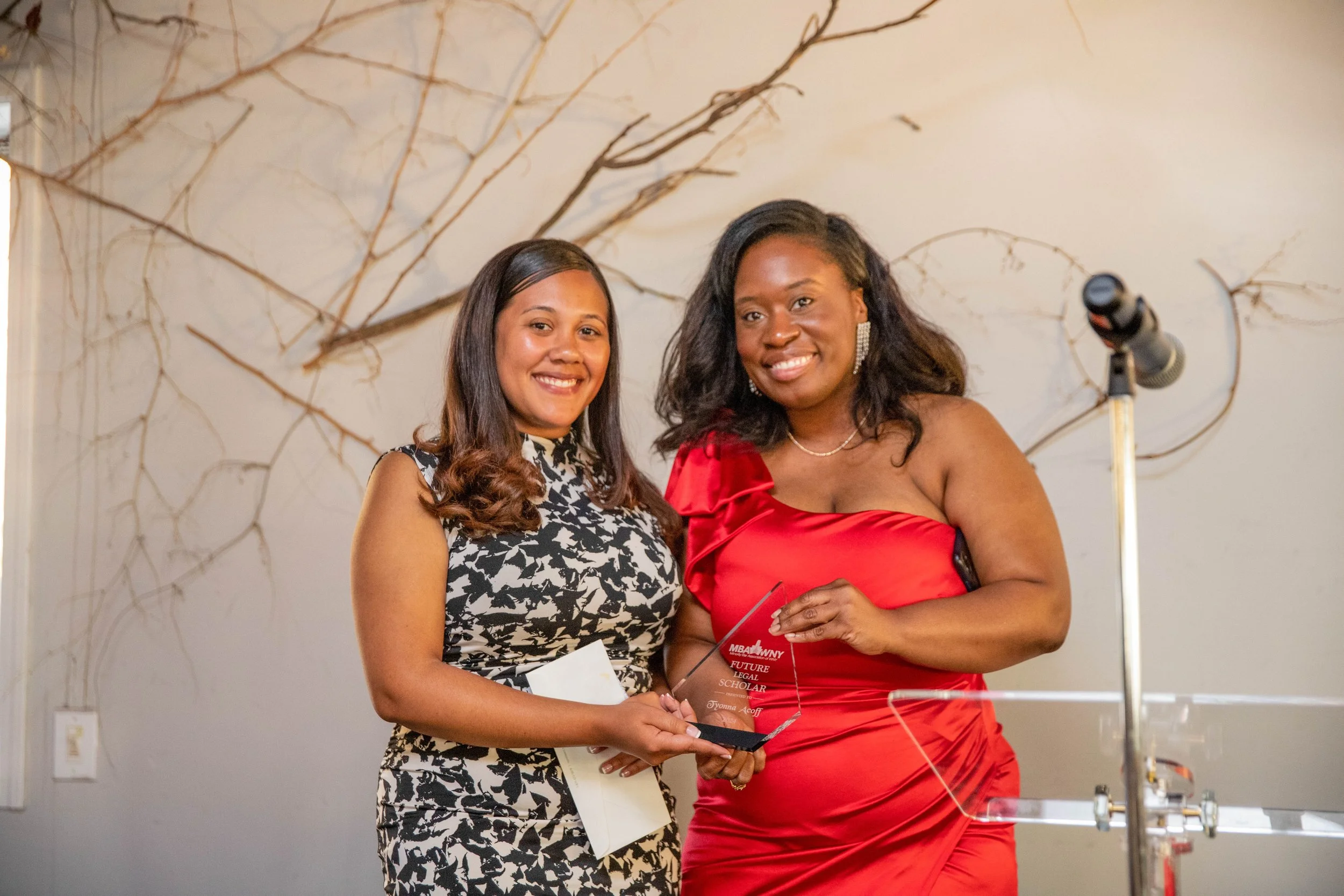 Two women standing together, one in a black and white patterned dress and the other in a bright red dress, smiling and holding an award or plaque. They are indoors with beige walls and decorative branches in the background.