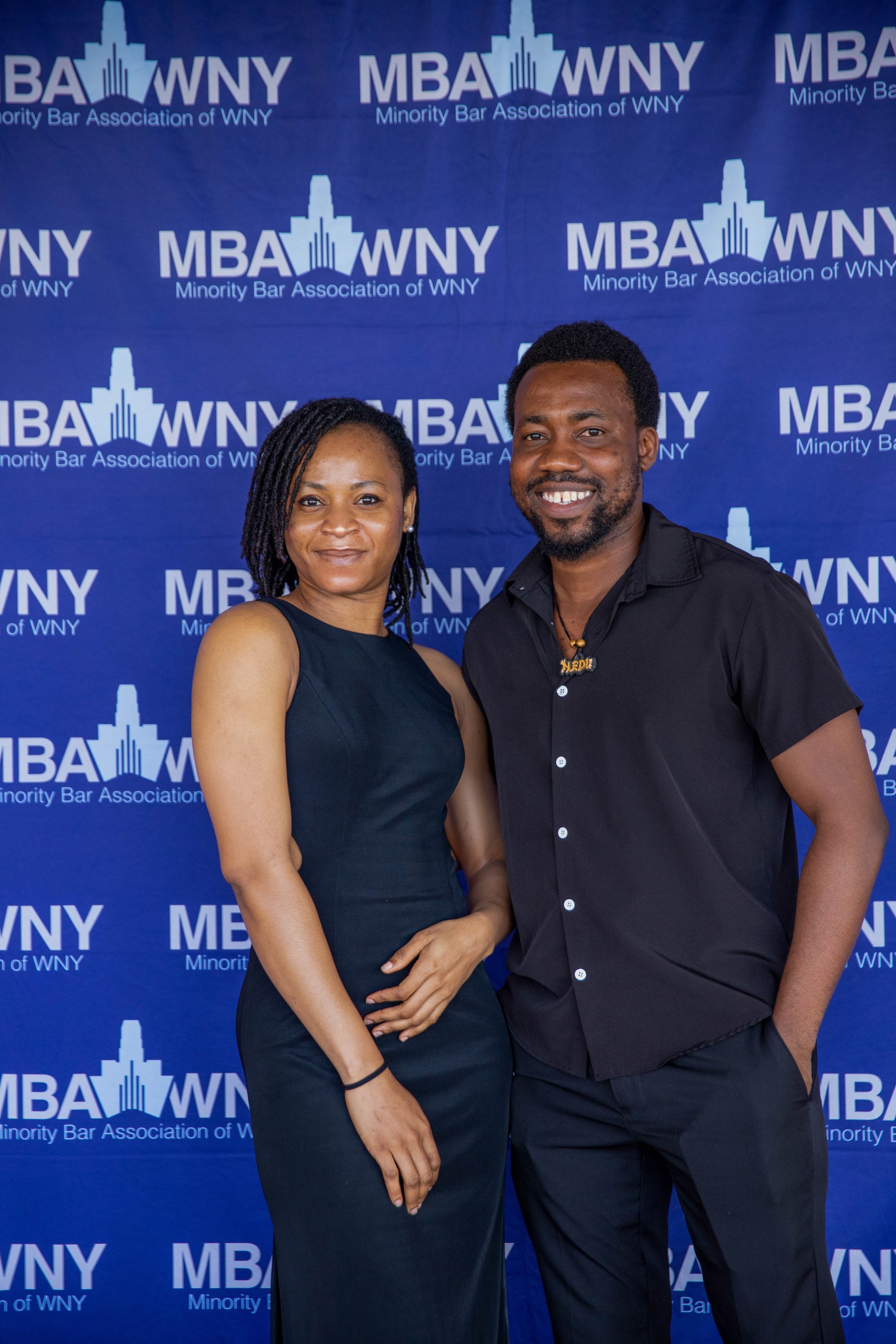 Two individuals, a woman with medium skin tone and a man with dark skin tone, standing in front of a blue backdrop with the logo and text for the Minority Bar Association of WNY. The woman is wearing a sleeveless black dress and has shoulder-length d