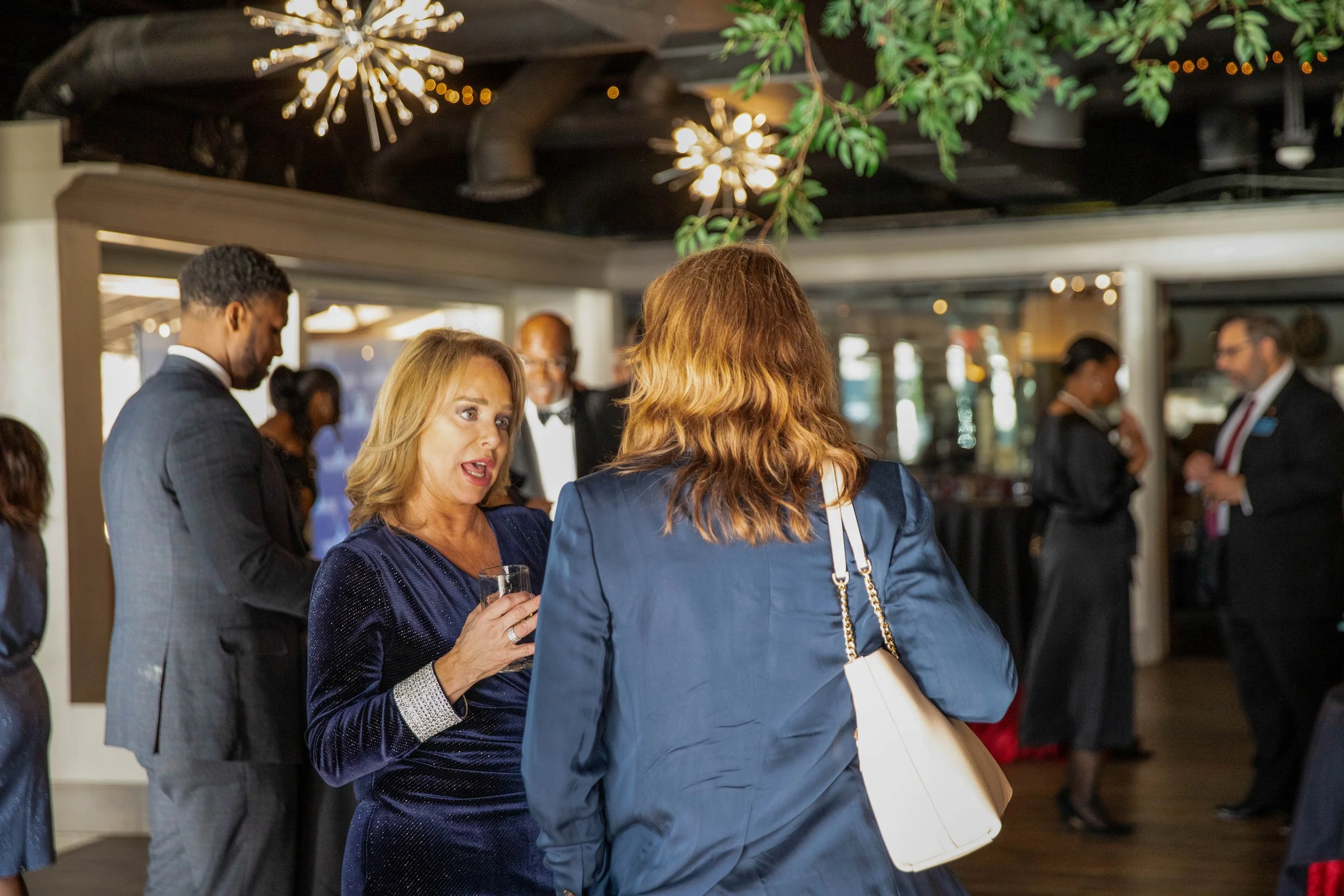 People dressed in formal attire socializing at an indoor event with decorative hanging lights and greenery.
