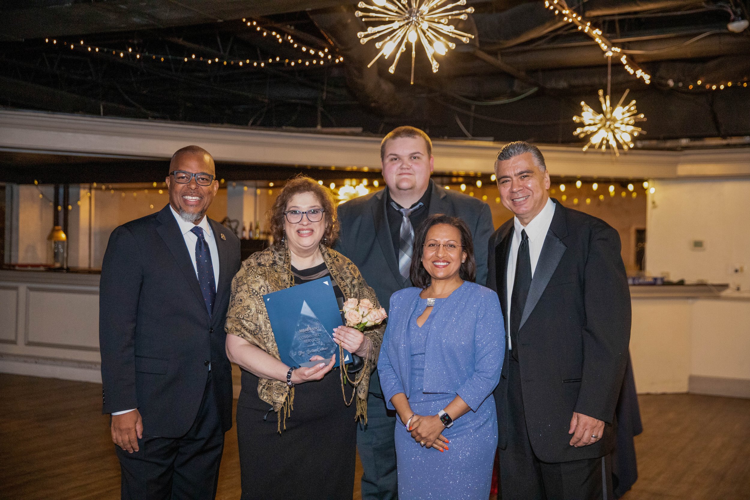 A group of five people, three men and two women, standing together indoors at a formal event. They are dressed in suits and a blue dress, smiling at the camera. The woman in the center holds a bouquet of pink roses and a glass award plaque. The backg