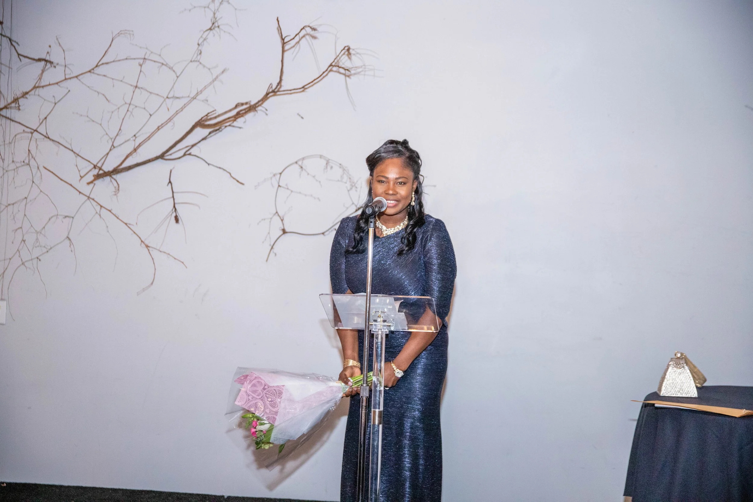 A woman dressed in a shiny, dark blue gown stands behind a transparent podium, speaking into a microphone, holding a bouquet of flowers wrapped in pink paper, with a wall decorated with a leafless tree branch in the background.