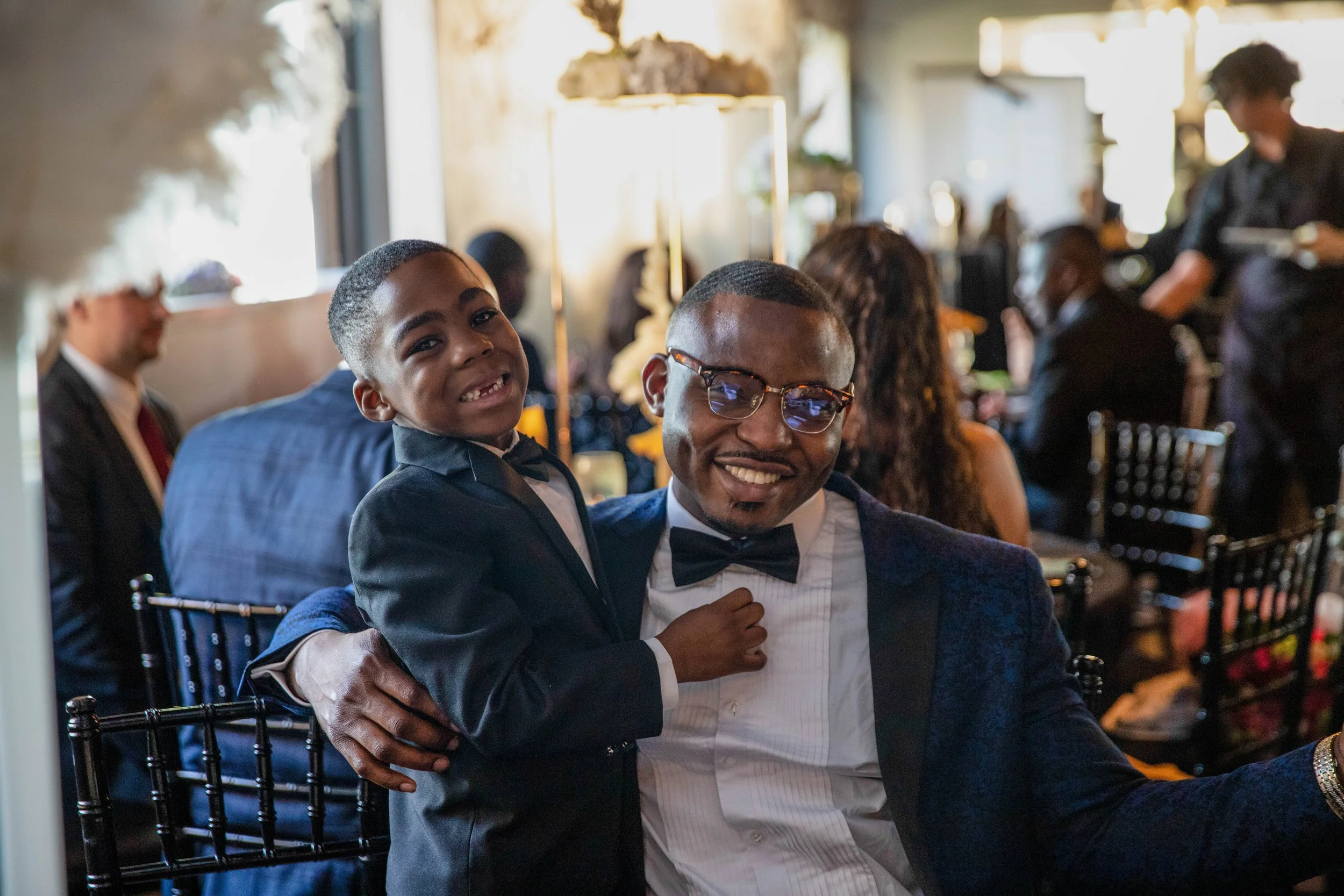 A man in a tuxedo and glasses cuddles a smiling young boy in a tuxedo, seated at a formal event with other guests and waitstaff in the background.