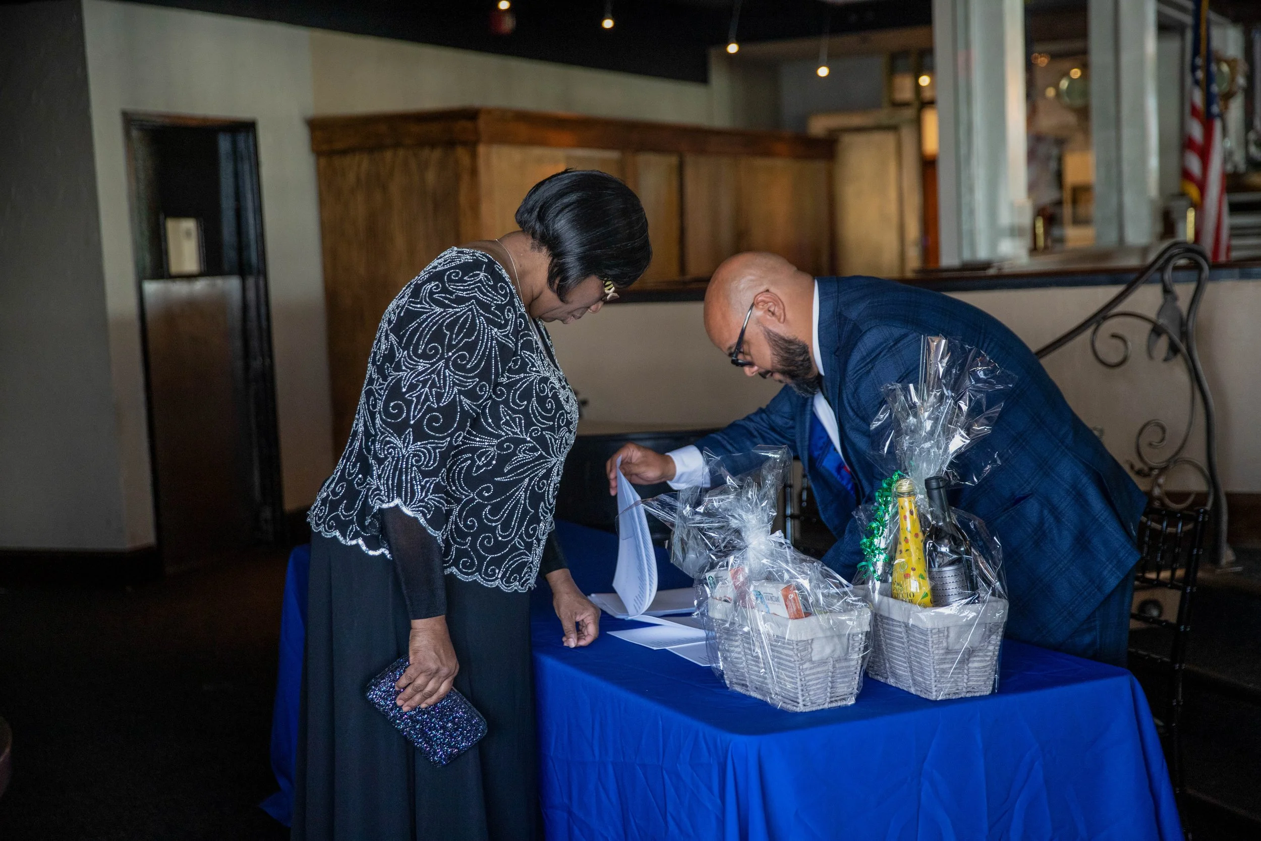 A man and a woman are bowing their heads at a table with gift baskets, possibly during a formal or celebratory event.