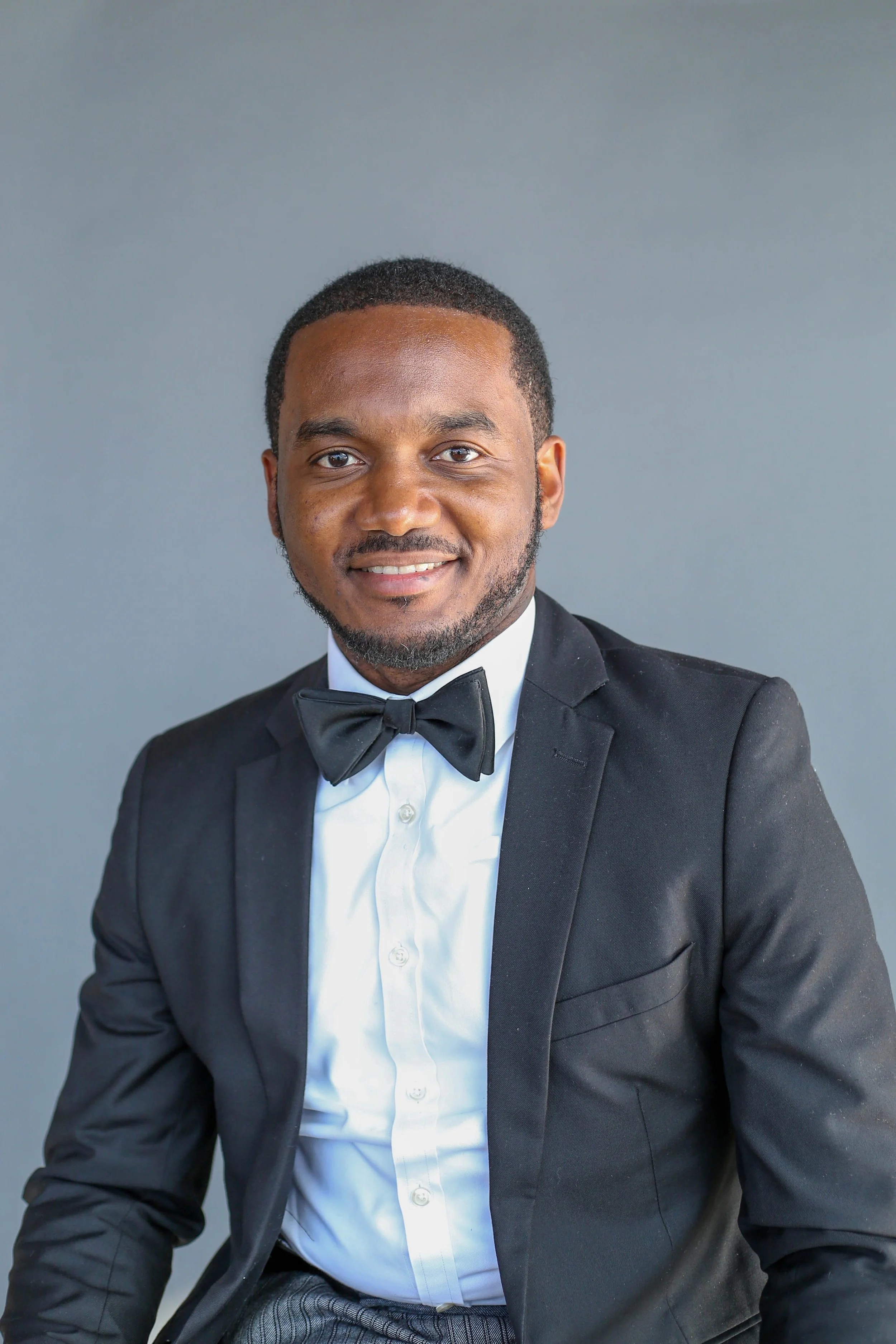 Portrait of a young man in a tuxedo, smiling, against a plain gray background.