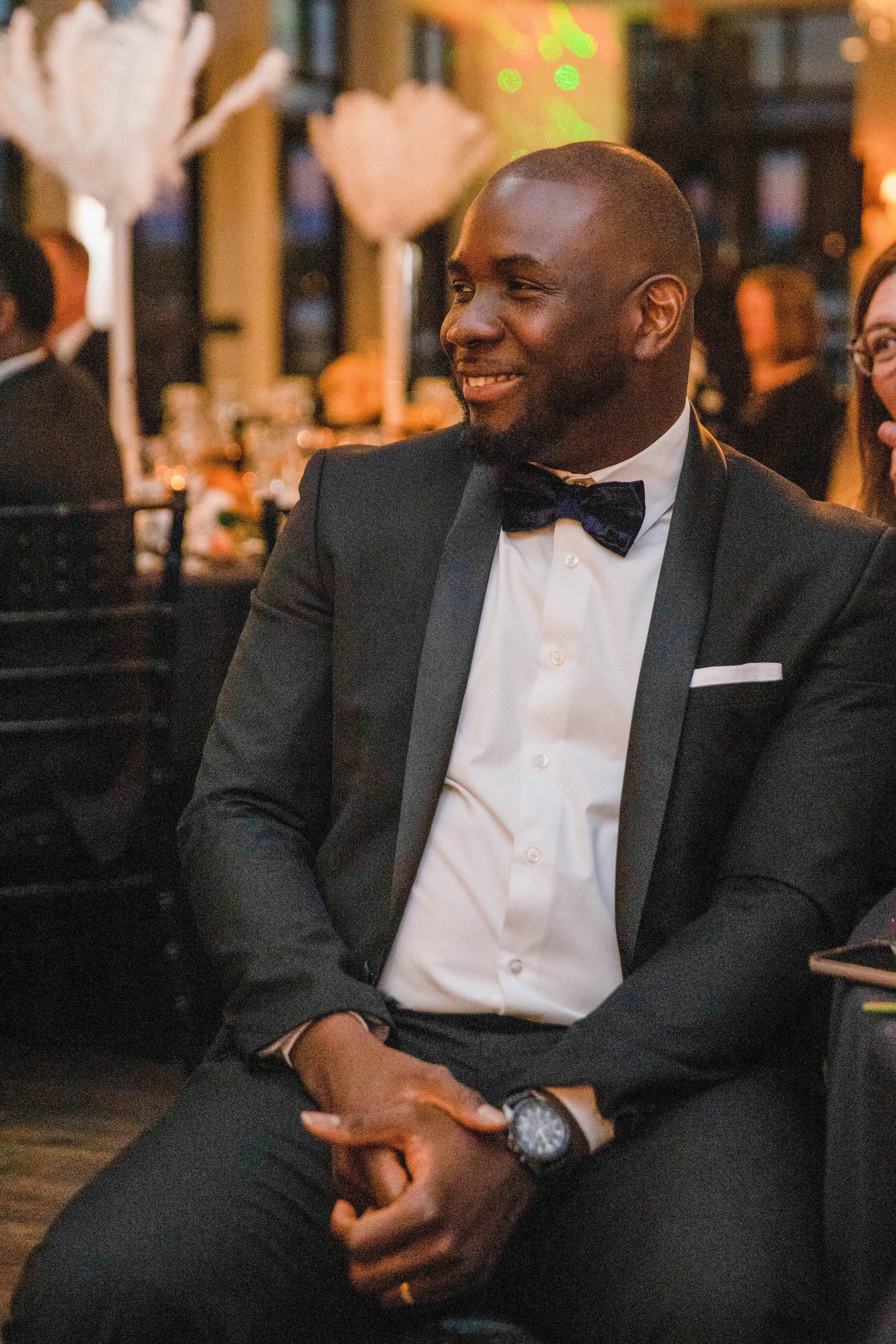 A man dressed in a tuxedo, smiling while sitting at a formal event, with a blurred background of other guests and decorative lighting.