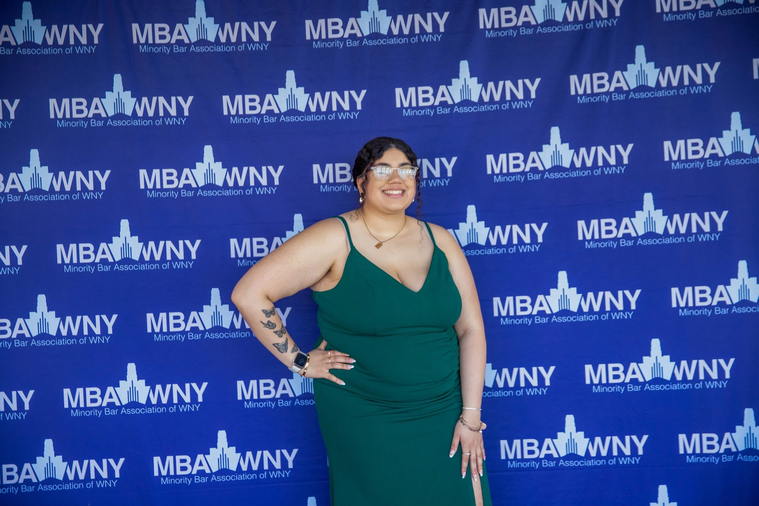 A woman in a green dress with tattoos on her arm, smiling, standing in front of a blue backdrop with the logo of the Minority Bar Association of WNY.
