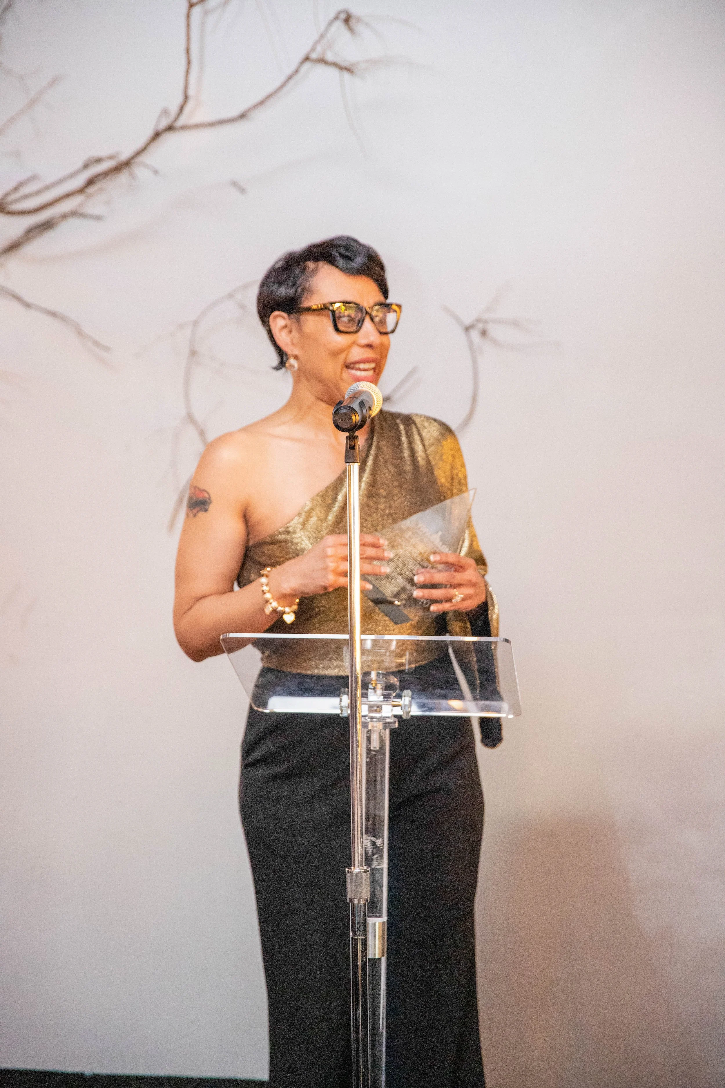 A woman with short black hair, glasses, and a tattoo on her left arm, wearing a gold and black dress, is speaking at a podium with a microphone.