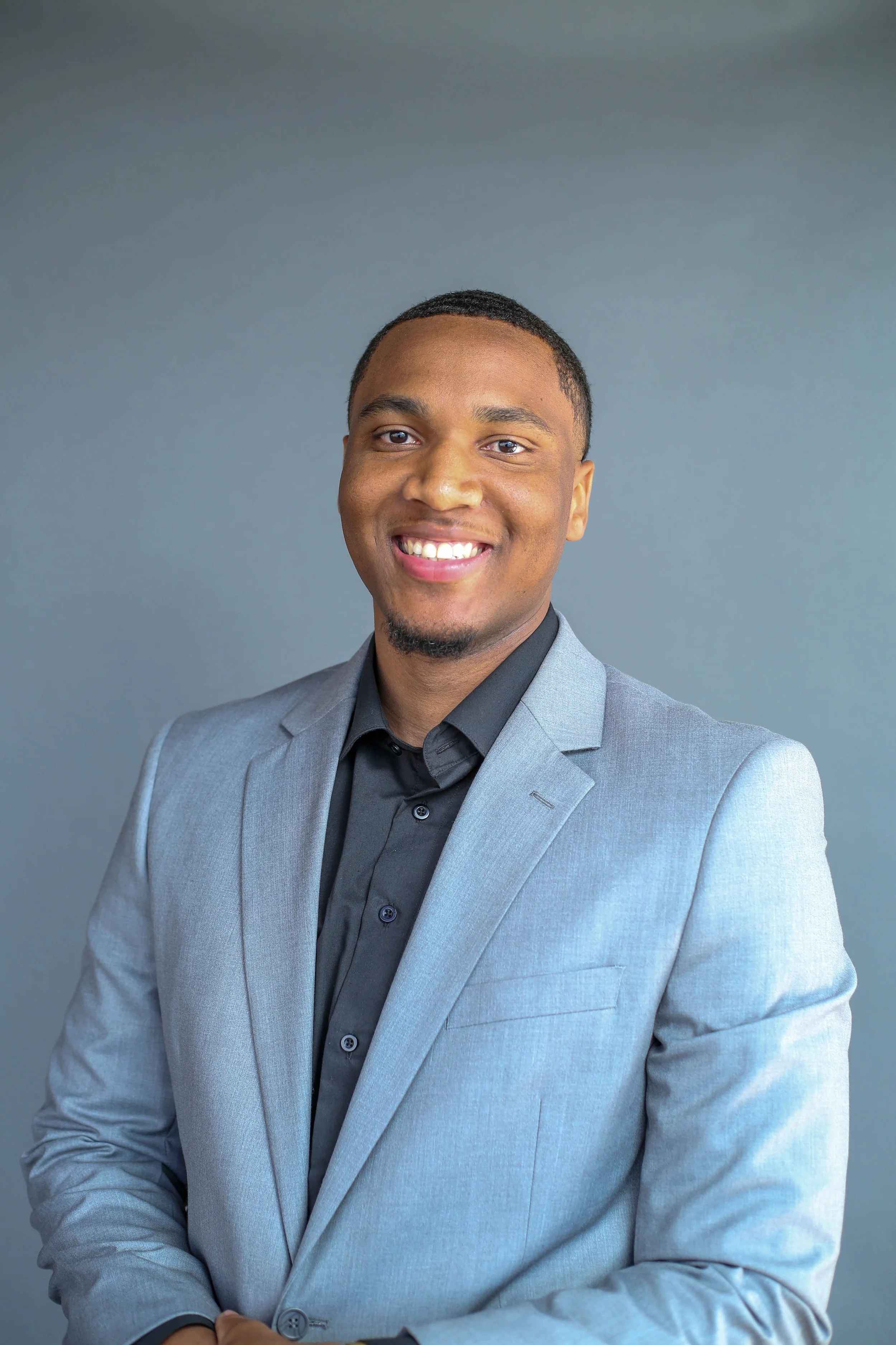 Young man smiling in a light gray suit and black shirt against a gray background.