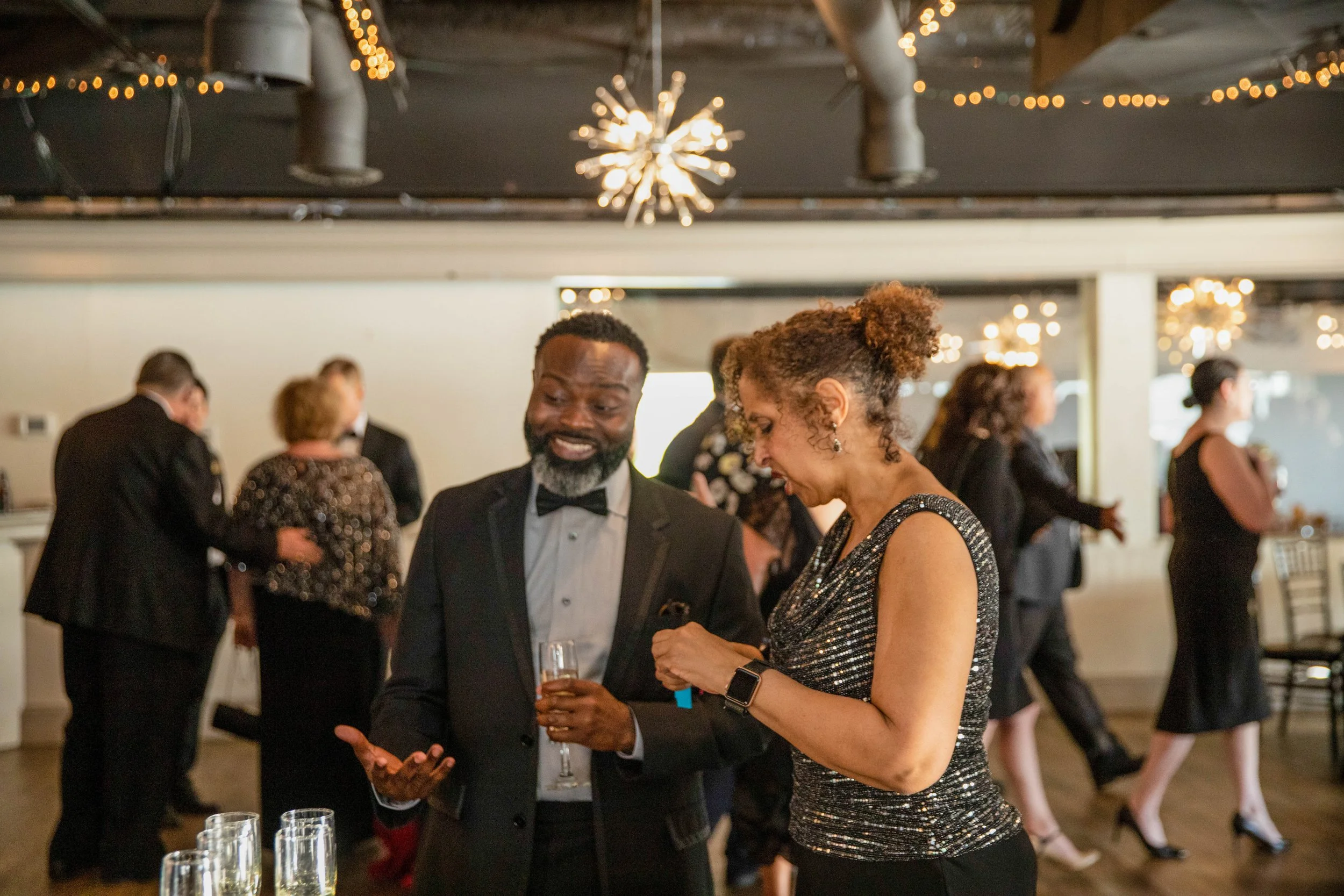 A couple dressed in formal attire dancing and smiling at a festive event with string lights and other guests in the background.