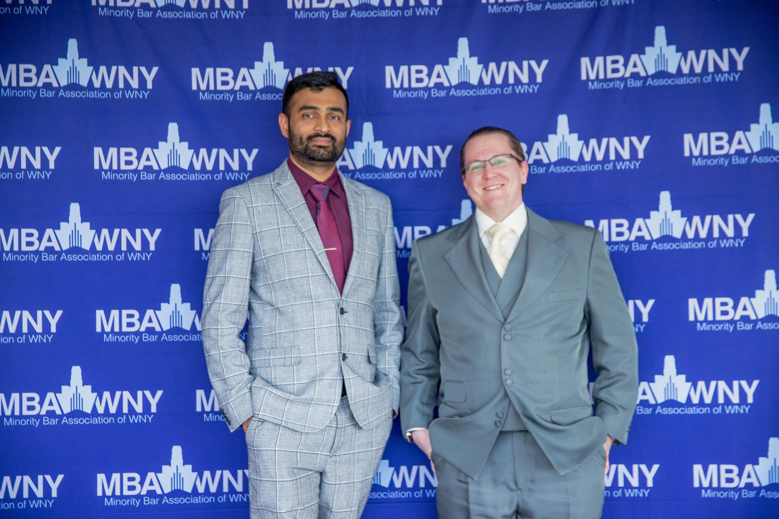 Two men in suits standing in front of a blue backdrop with the logo and text of the Minority Bar Association of WNY.