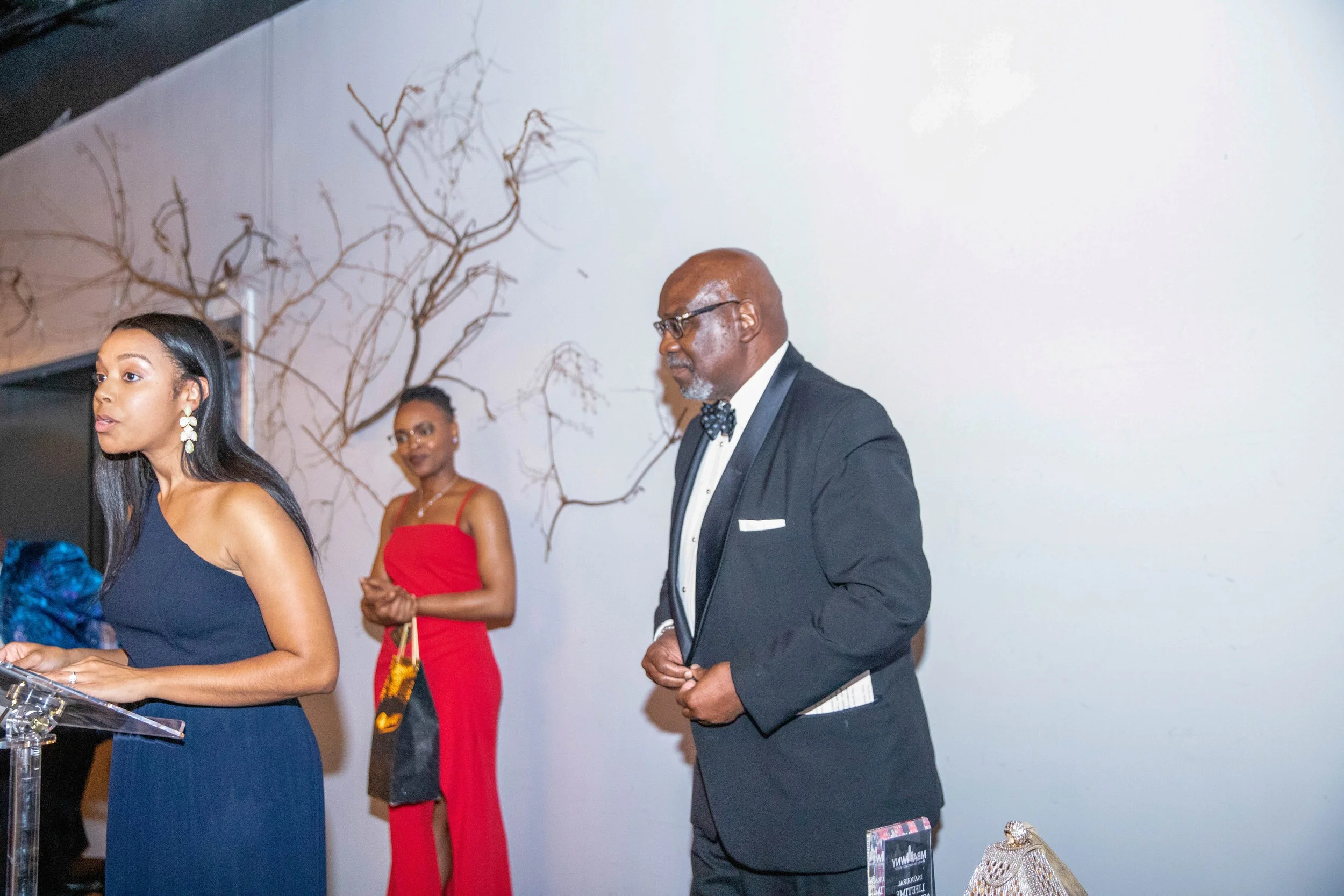 Three people at an indoor event dressed formally; women in elegant dresses, man in a tuxedo, standing in front of a white wall with a decorative branch sculpture.