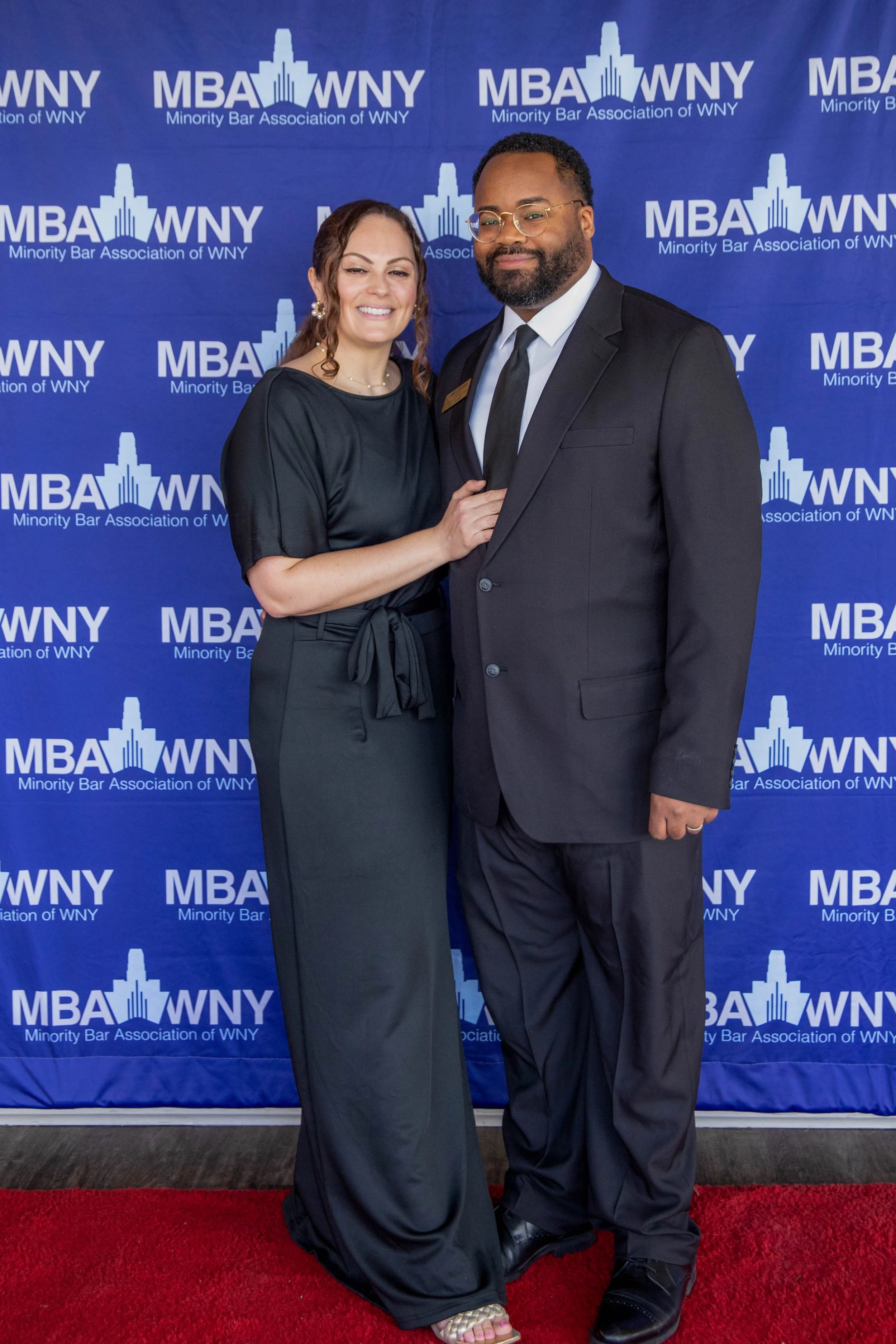 A woman and a man dressed formally stand on a red carpet in front of a blue backdrop with the logos of the Minority Bar Association of WNY. The woman is smiling, has long wavy brown hair, and wears a black dress, pearl earrings, and sandals. The man,