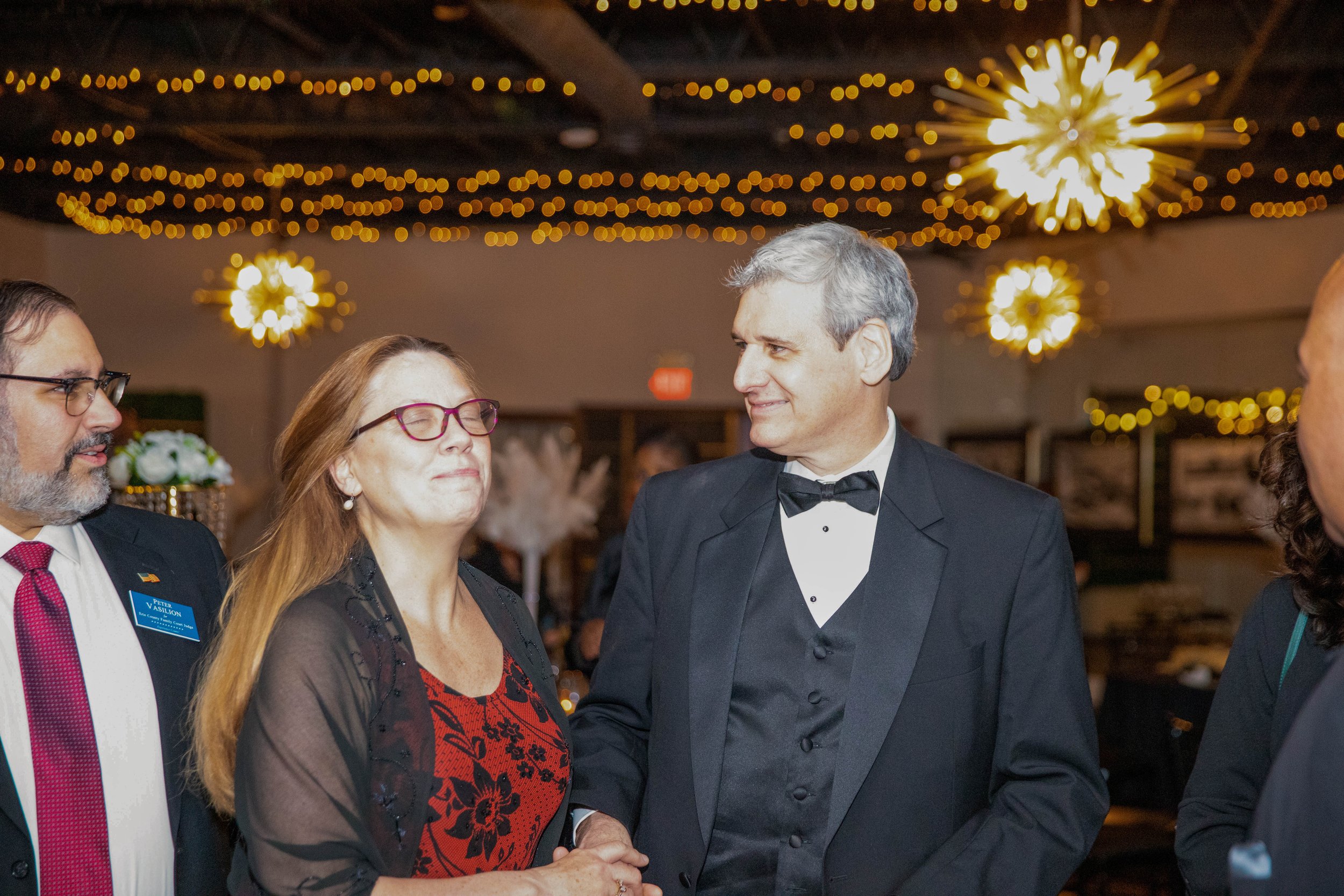 Four people in formal attire having a conversation at a fancy event with warm lighting and decorative ceiling lights.