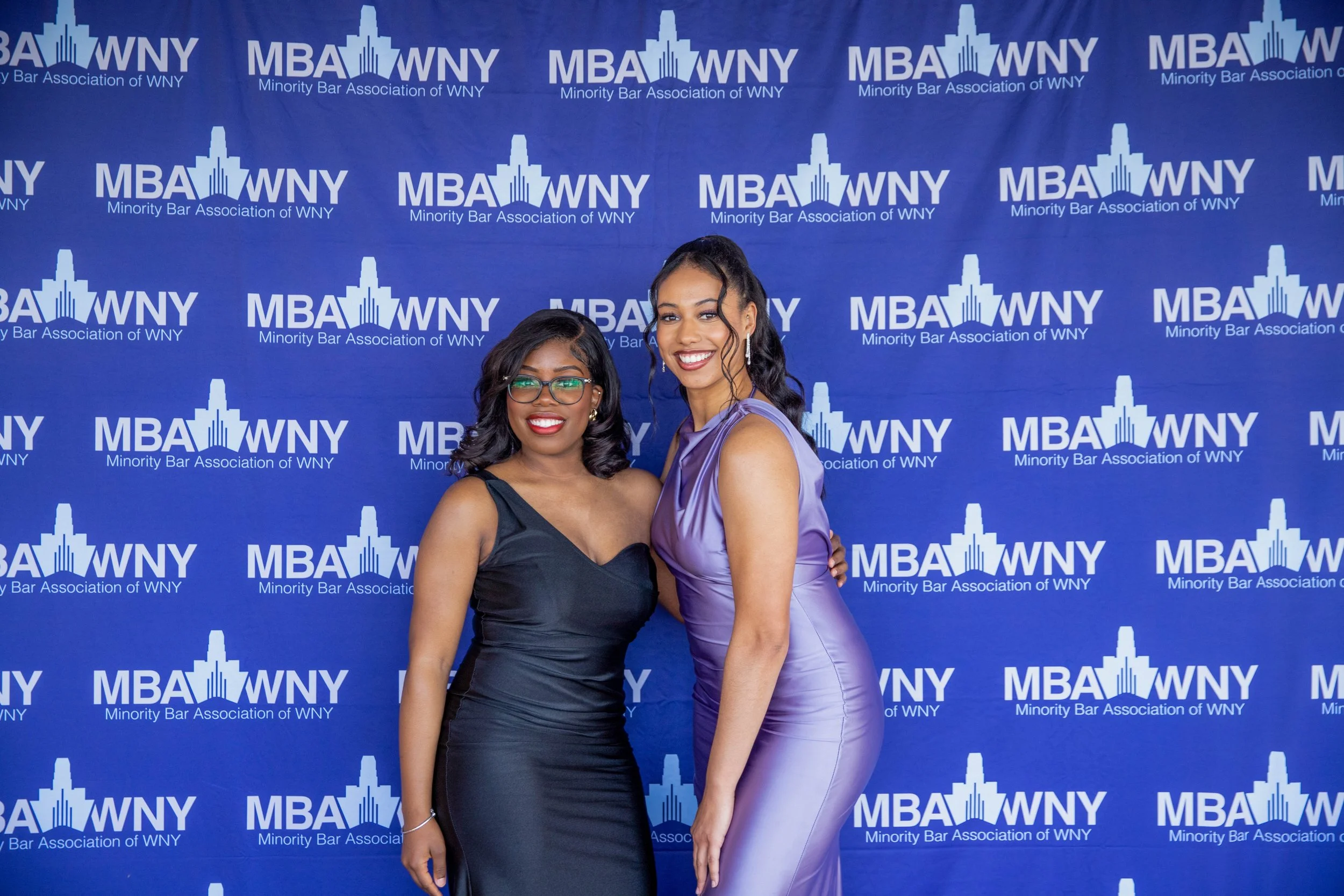 Two women dressed in formal attire posing in front of a blue backdrop with the logos of the Minority Bar Association of WNY. The woman on the left is wearing a black satin dress and glasses, and the woman on the right is wearing a lavender satin dres