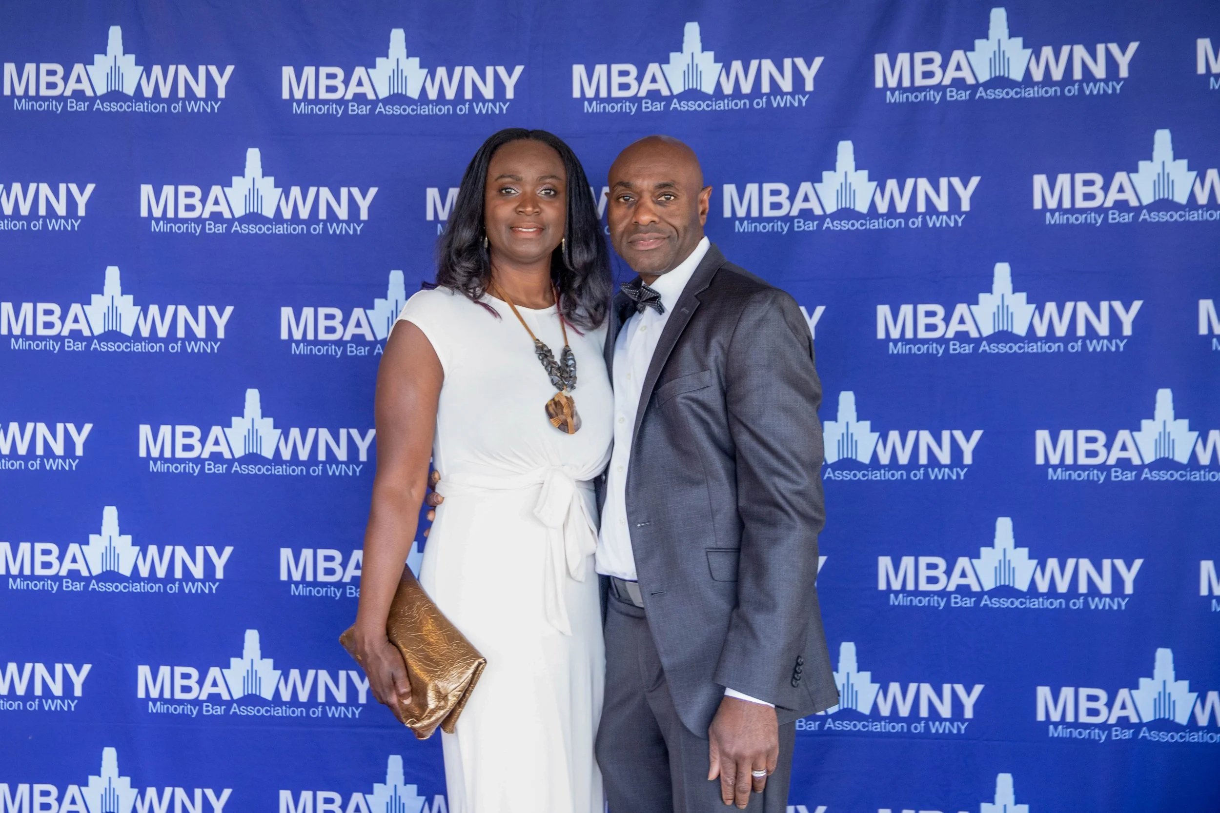 A couple dressed in formal attire standing in front of a blue backdrop with the logo and text 'MBA WNY Minority Bar Association of WNY.' The woman is wearing a white dress, a large necklace, and is holding a gold clutch. The man is dressed in a dark 