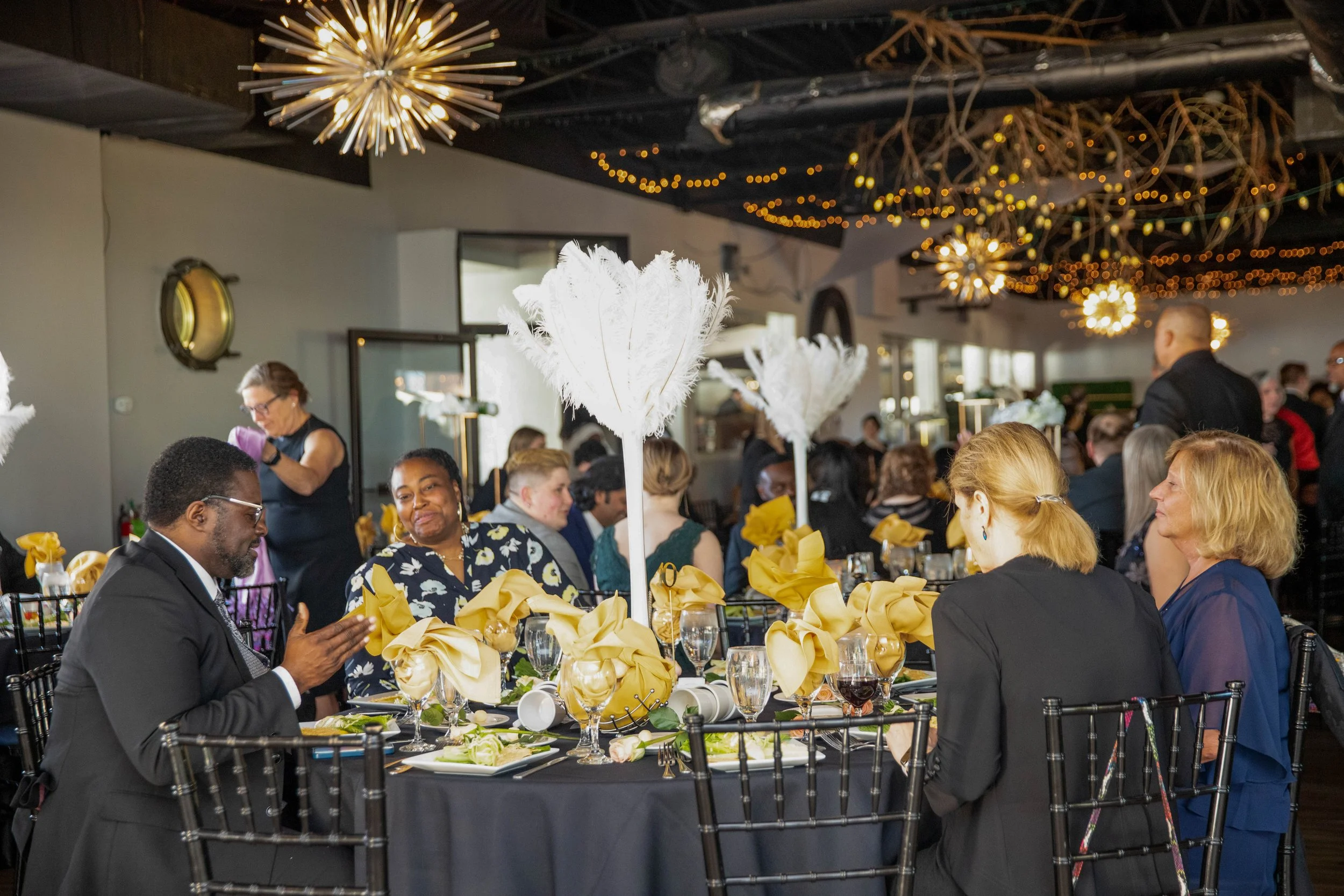 A crowded indoor event with people seated at decorated tables, white feather centerpieces, and warm string lights hanging from the ceiling.