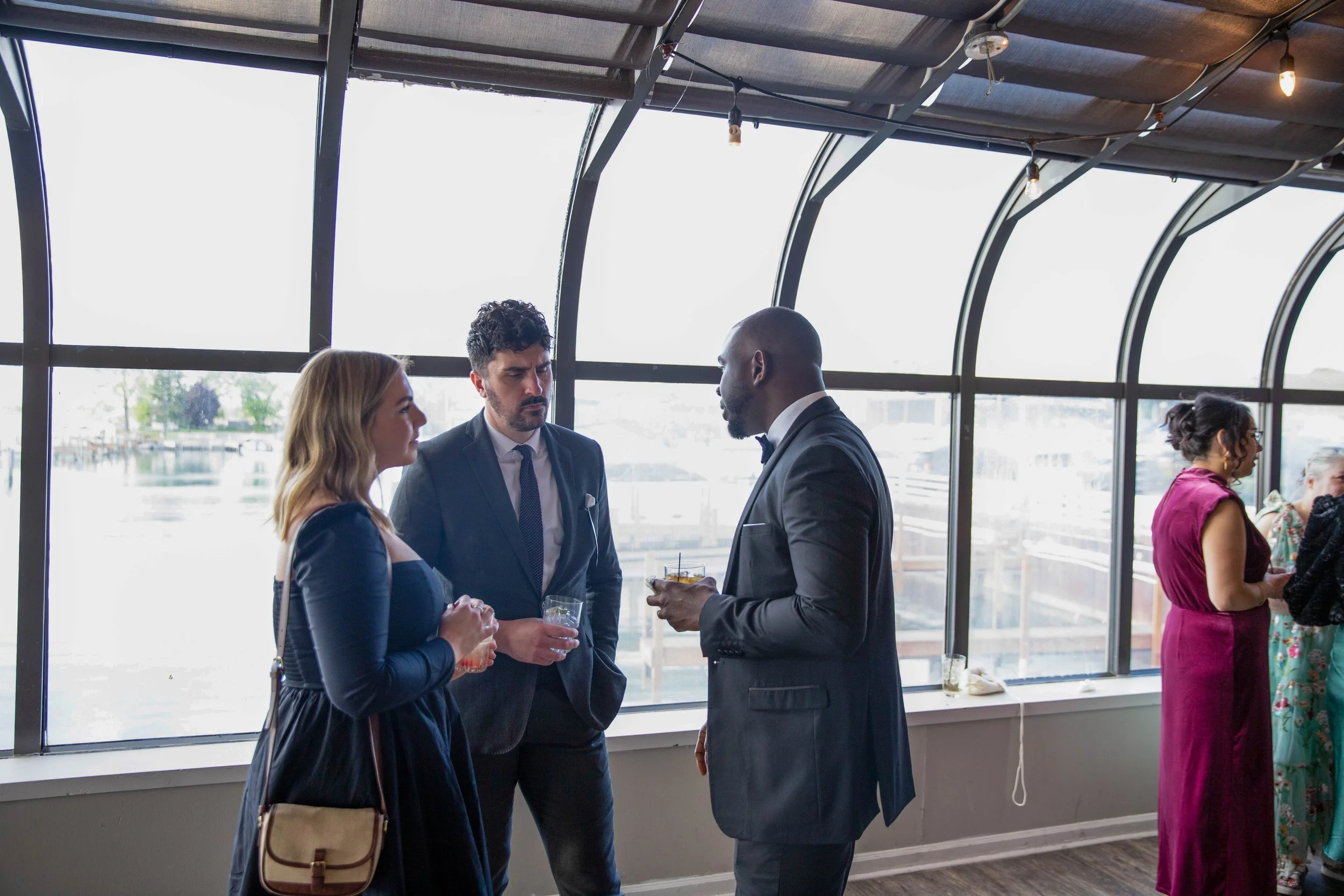 Three people dressed in formal attire are engaged in conversation at a social event indoors near large windows with a view of a marina or waterfront. Two women and one man, all holding drinks, are talking, while others are visible in the background.
