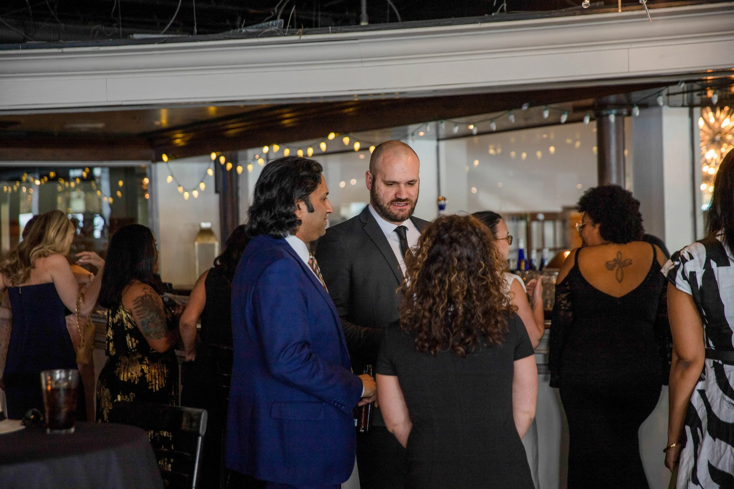 A group of people socializing at a party in a dimly lit venue. Some are dressed in formal attire, and there are string lights hanging overhead.