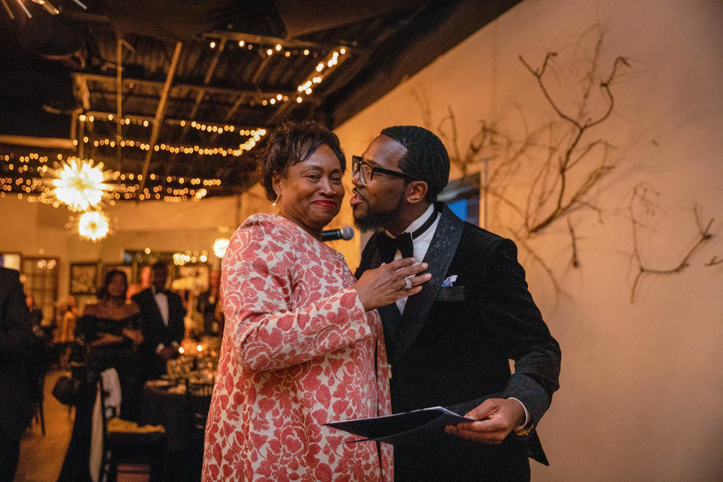 An older woman and a man are dancing closely at a formal event, with background guests and warm, decorative lighting.
