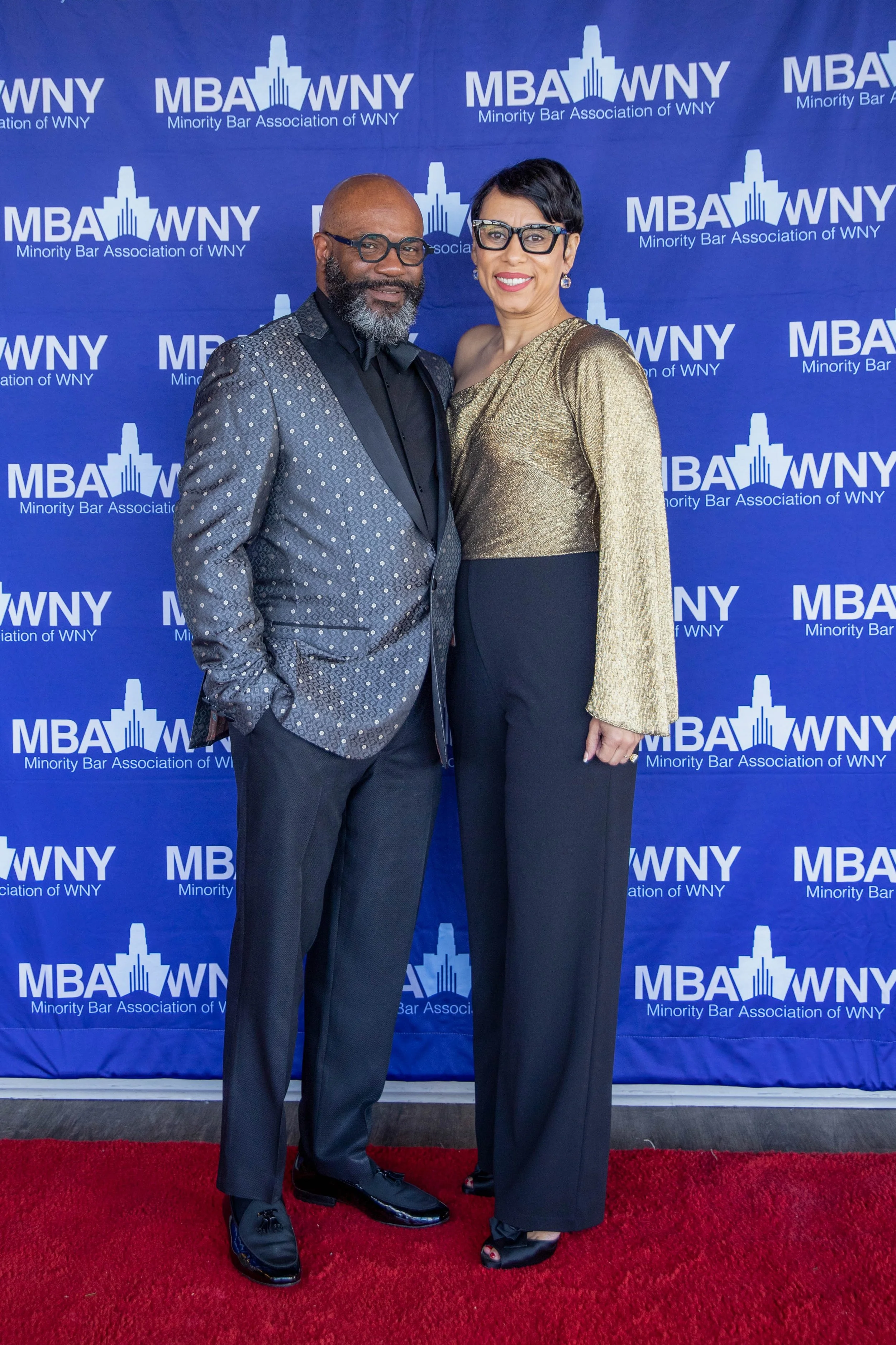 Two people standing on a red carpet in front of a blue backdrop with the logo and text 'Minority Bar Association of WNY'. The man on the left is wearing a patterned tuxedo with a black shirt and black shoes, and has a bald head with a gray beard. The