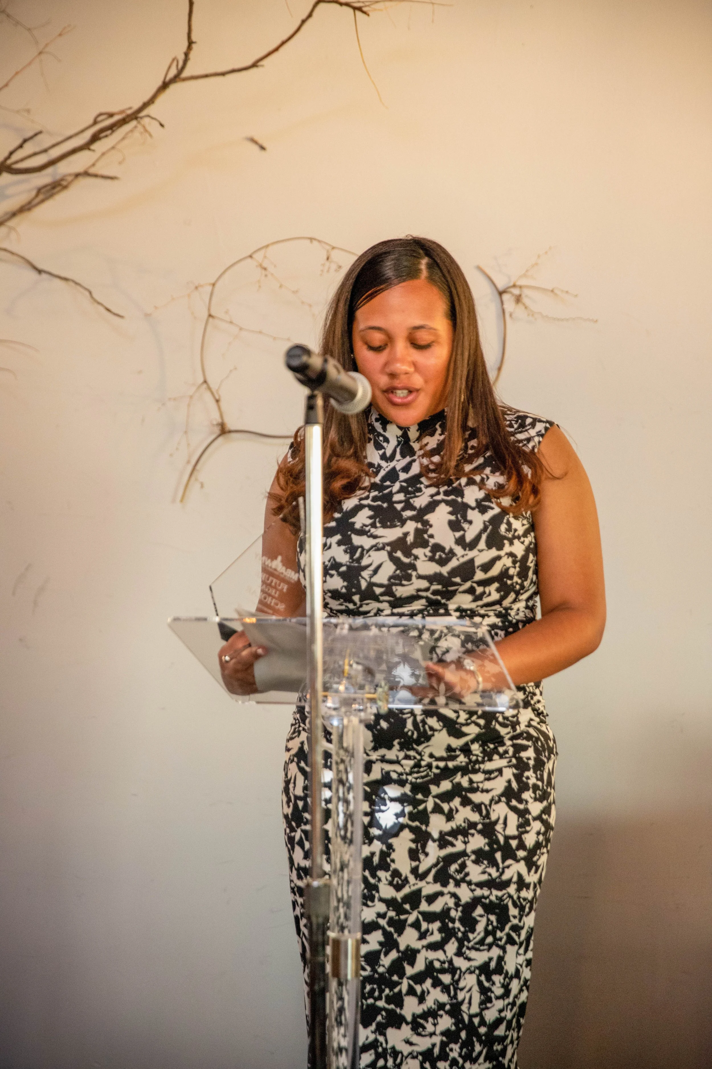 A woman in a black and white patterned dress stands at a transparent podium, reading from a paper, with a microphone in front of her. A decorative branch is seen on the wall behind her.