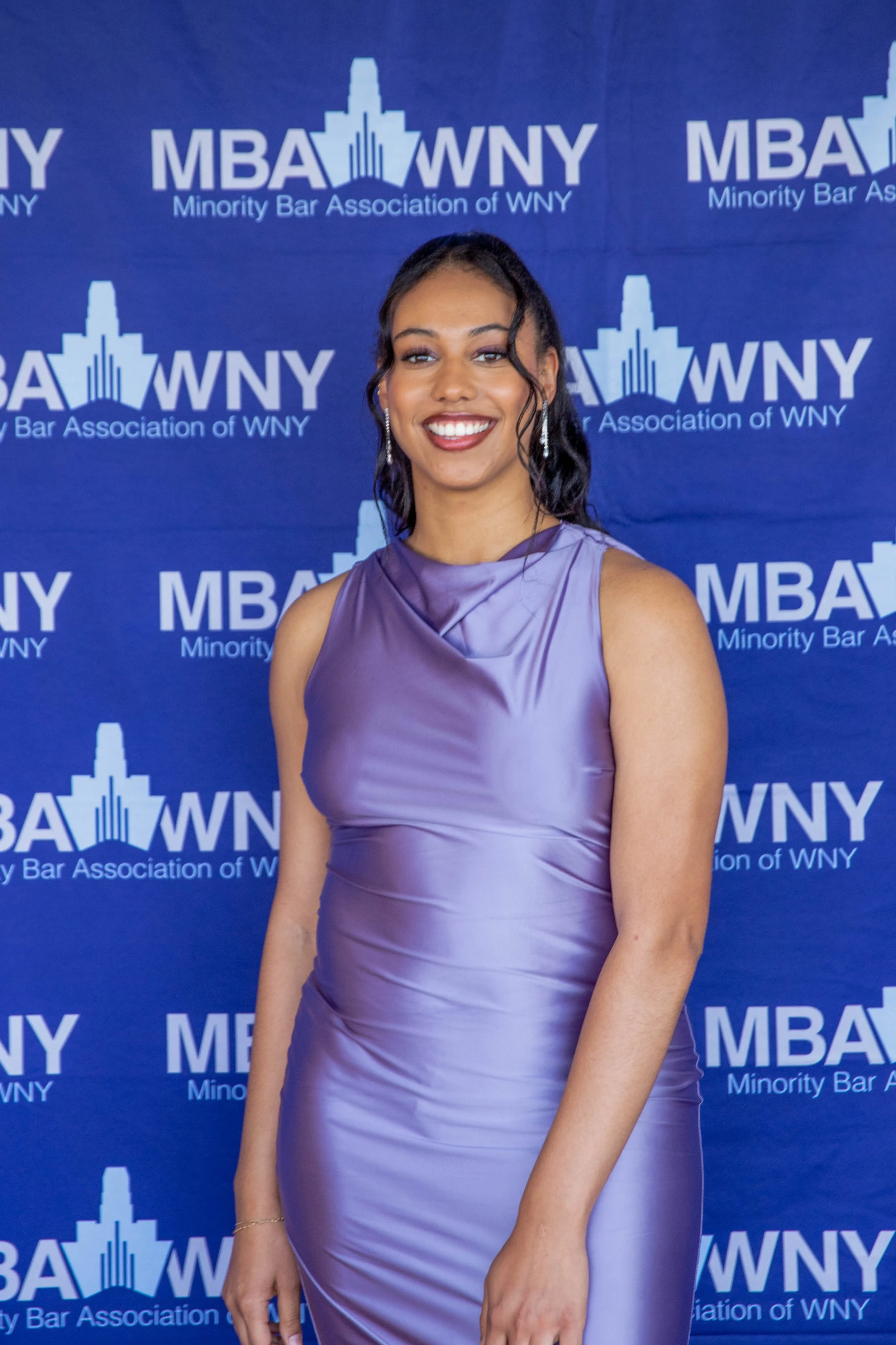 A woman in a lavender satin dress smiling, standing against a blue backdrop with the logo of the Minority Bar Association of WNY.
