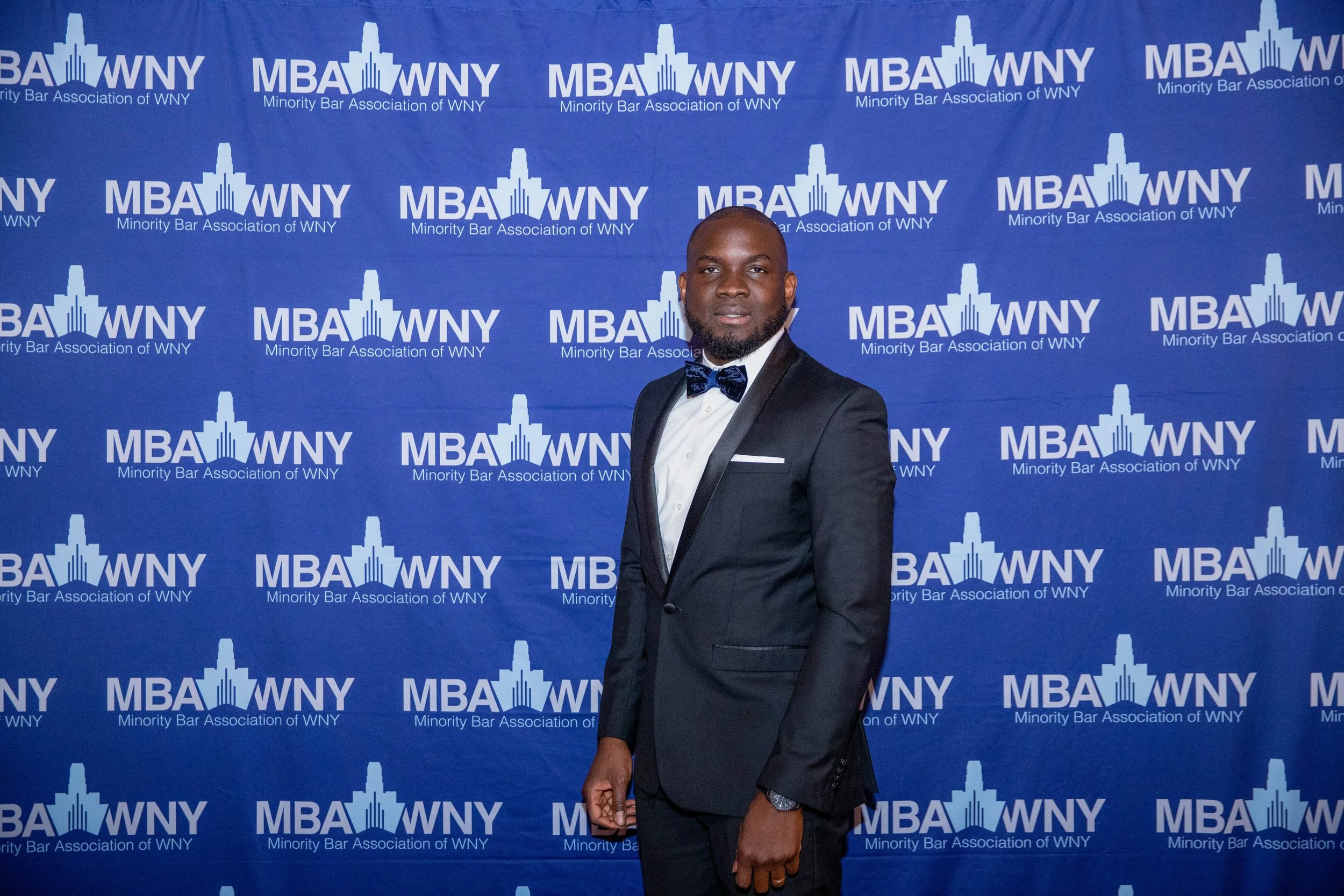 A man in a tuxedo with a bow tie standing in front of a blue backdrop with the logo and text for the Minority Bar Association of WNY.