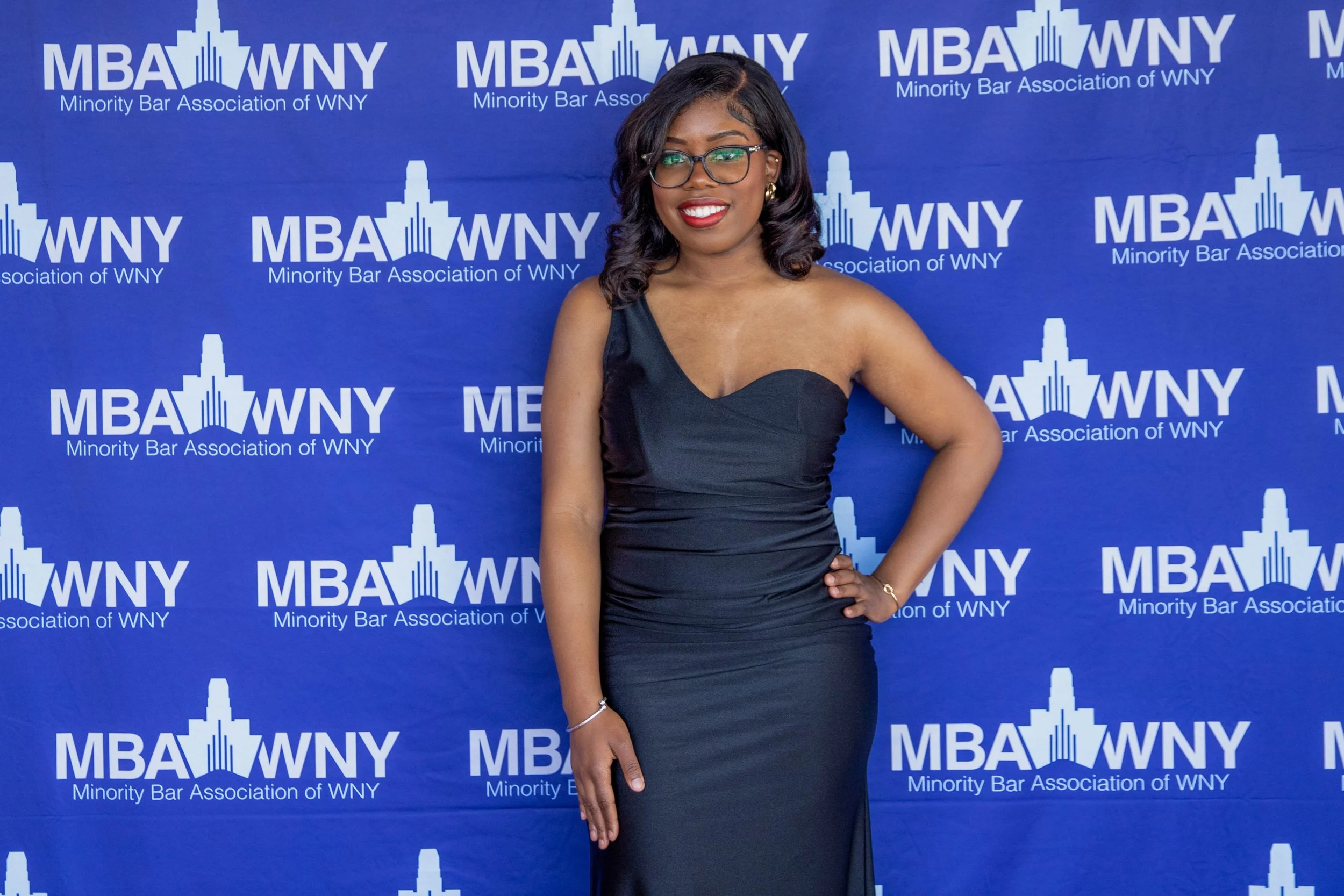 A woman in a black evening gown and glasses posing in front of a blue backdrop with 'Minority Bar Association of WNY' and 'MBA WNY' logos.