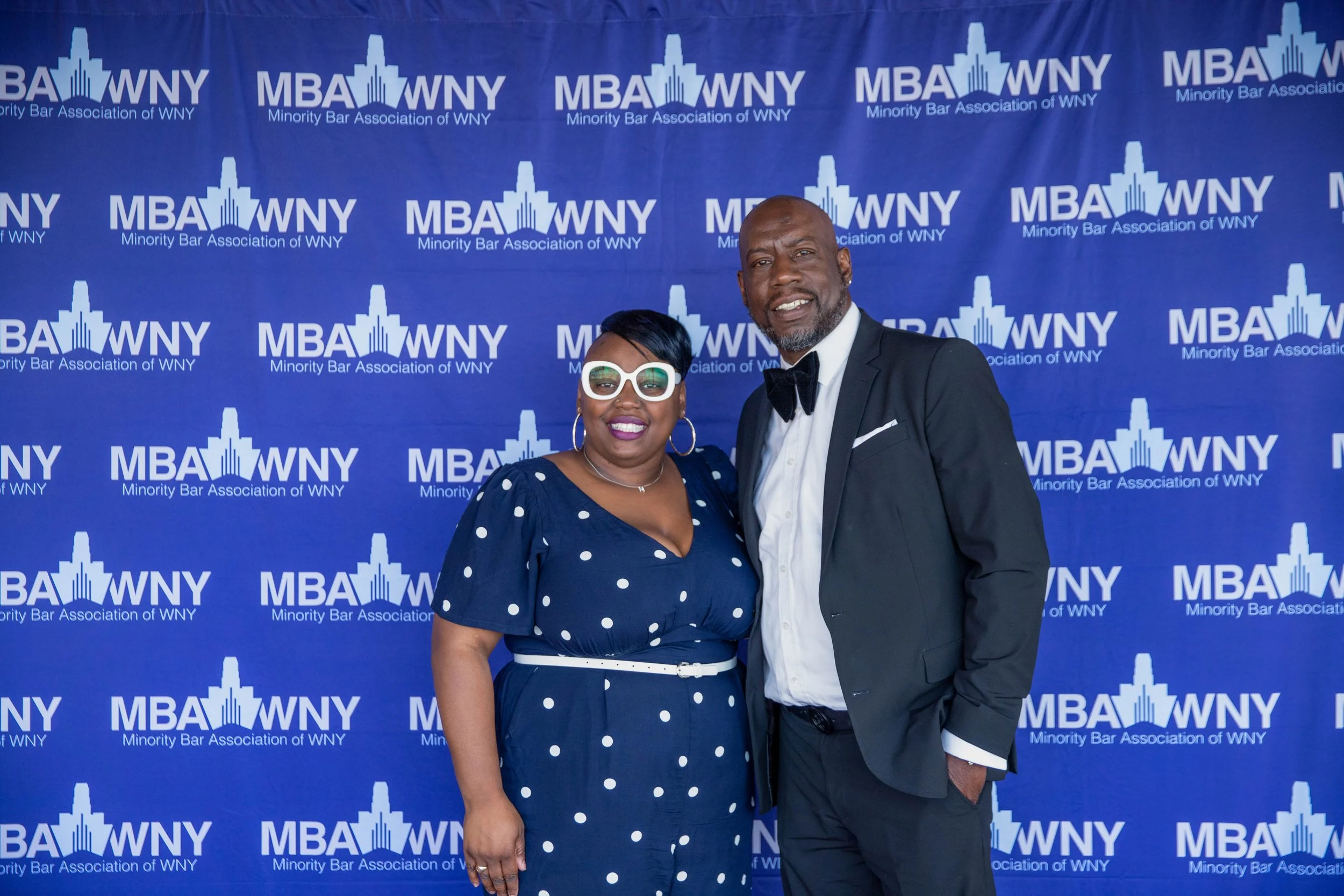 Two people standing in front of a blue backdrop with the text 'MBA WNY Minority Bar Association of WNY' repeated across it. The woman on the left is wearing a navy blue dress with white polka dots, white sunglasses, and hoop earrings. The man on the 