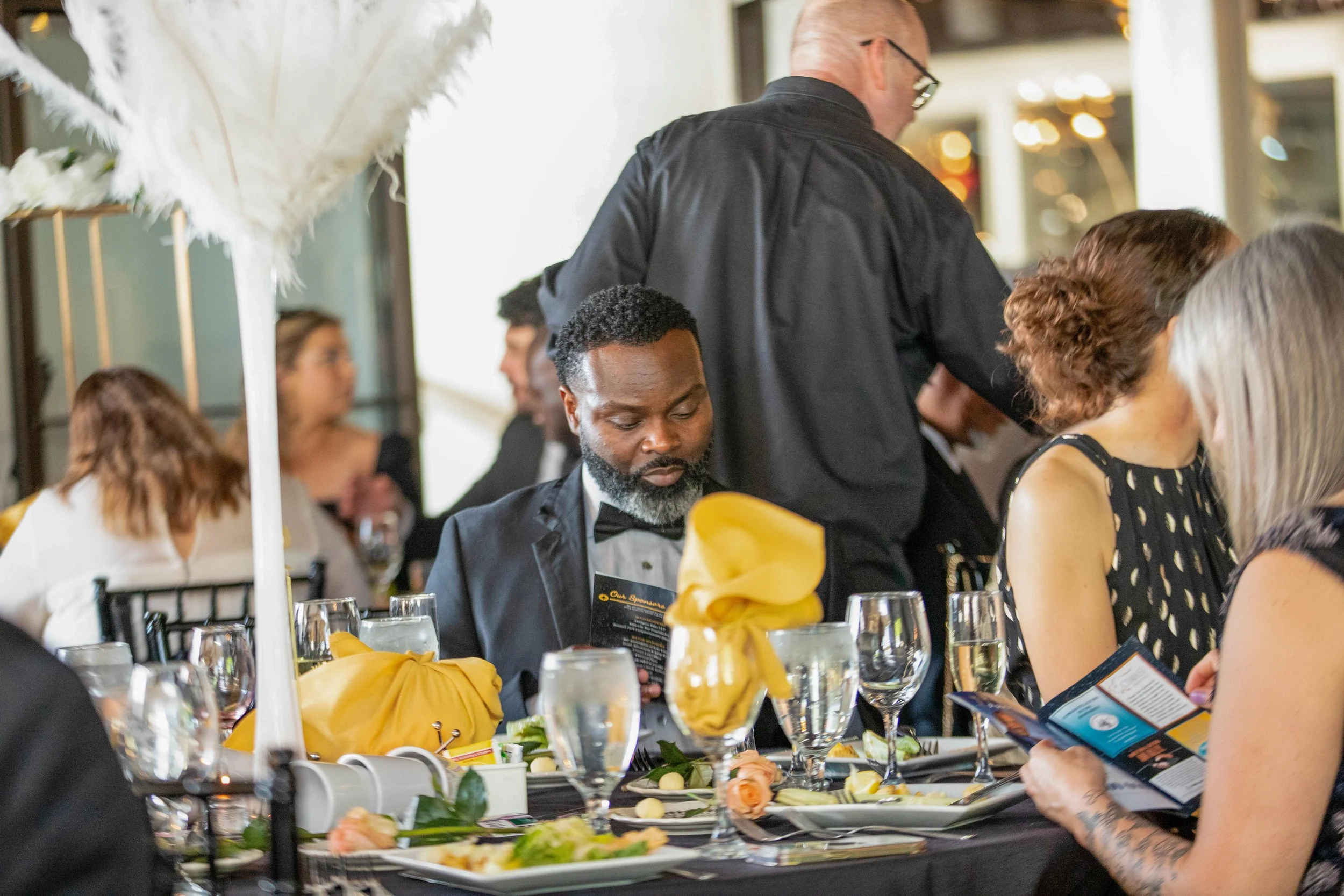 People seated at a formal event, with one person in focus wearing a tuxedo and bouquet of flowers on the table in front of him, others standing and dressed formally, set at a decorated dining table with glasses, plates, and yellow napkins.