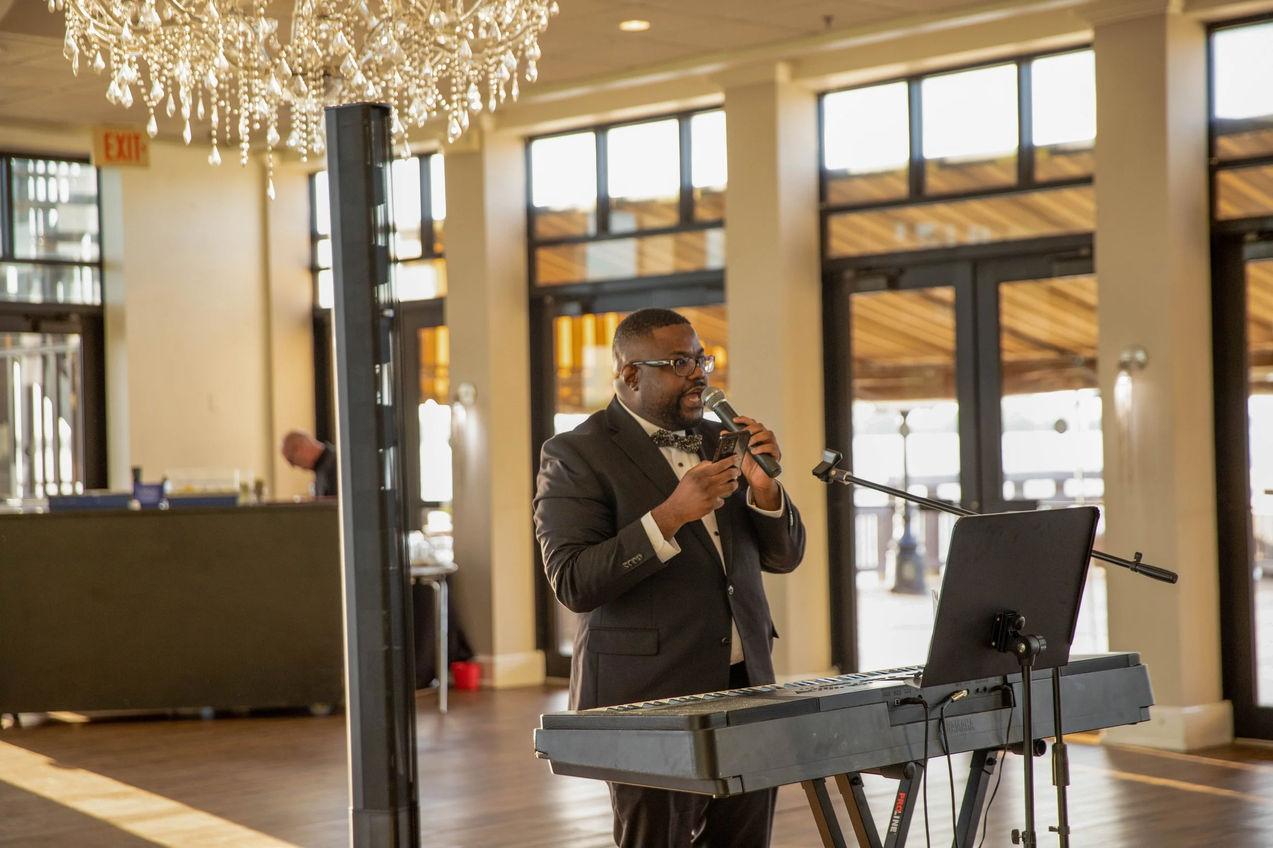 A man in a tuxedo with glasses is playing a keyboard and singing into a microphone in a well-lit indoor space with large windows and a chandelier hanging from the ceiling.