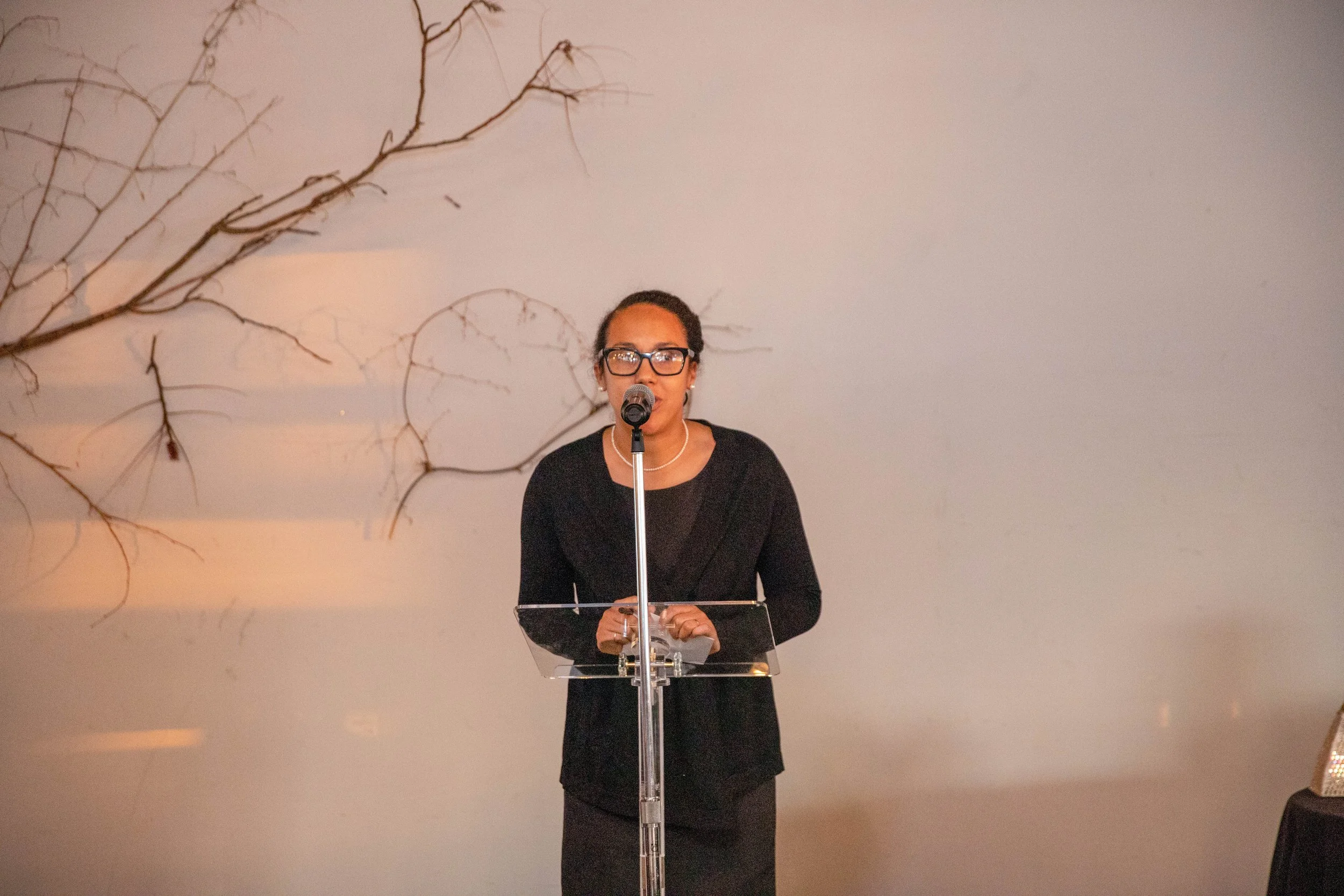 A woman wearing glasses, a black top, and a pearl necklace stands behind a clear podium speaking into a microphone against a plain background with a large, leafless branch art piece on the wall.