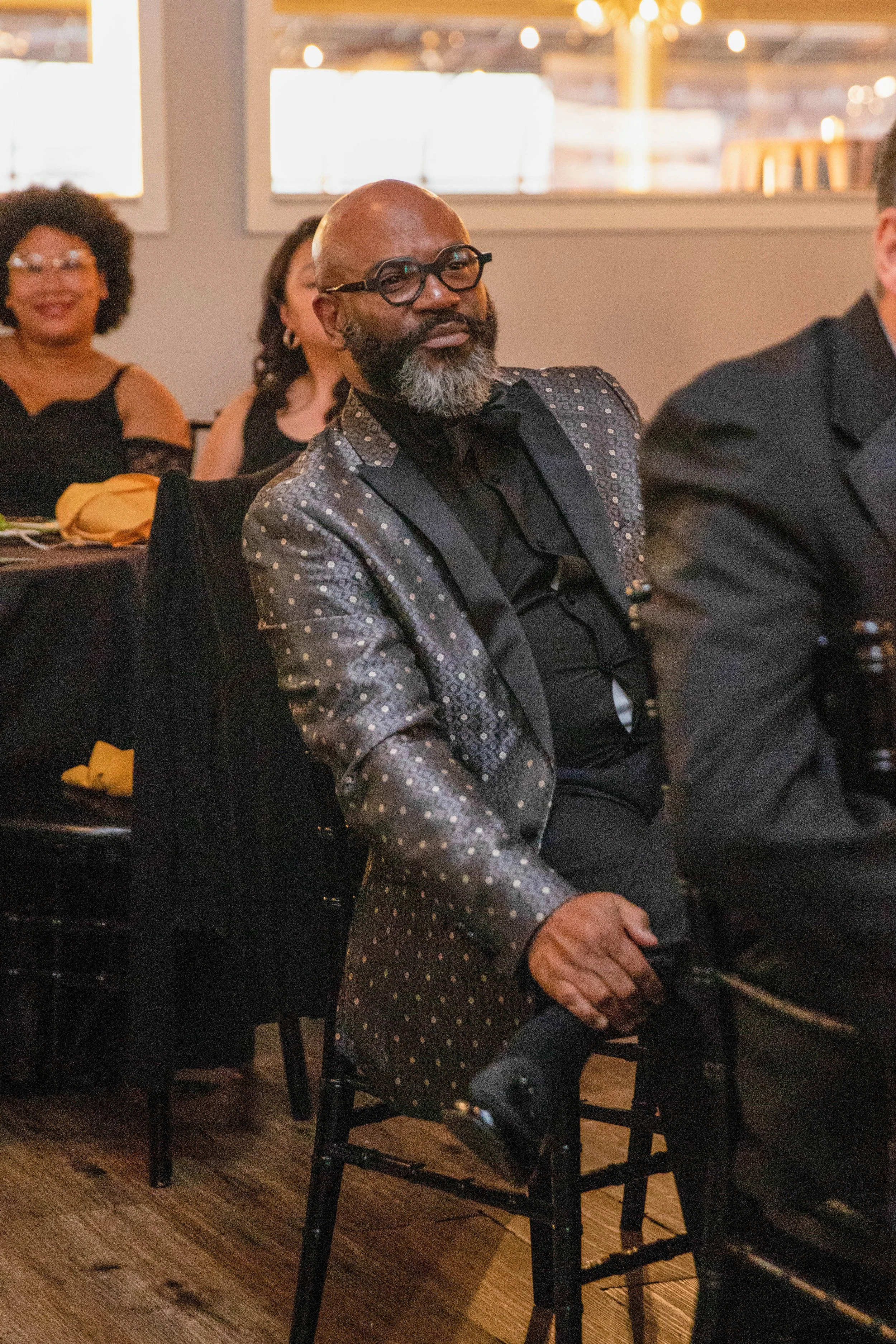 A man with a bald head, glasses, and a beard, dressed in a patterned blazer and black shirt, sitting at a table during an indoor event. Several women are seated behind him, smiling and wearing stylish black outfits. The setting appears to be a restau