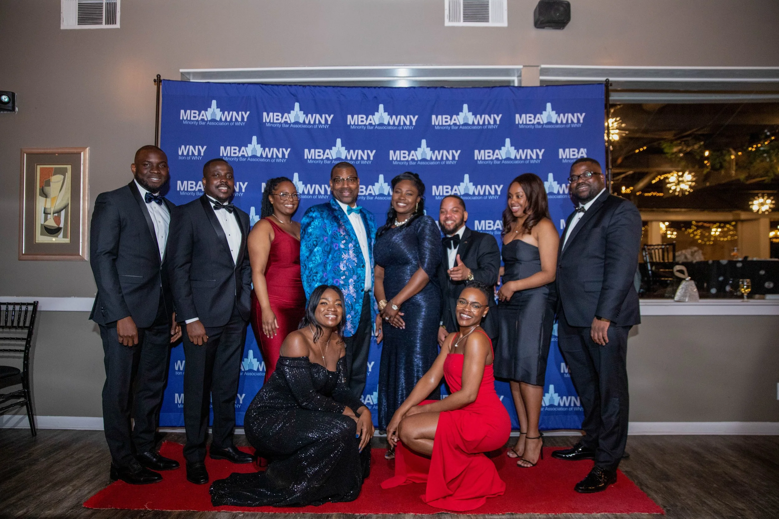 Group of nine people dressed formally at an event, standing in front of a blue backdrop with the logo 'MBA WNY,' on a red carpet, with some sitting in front and others standing behind.