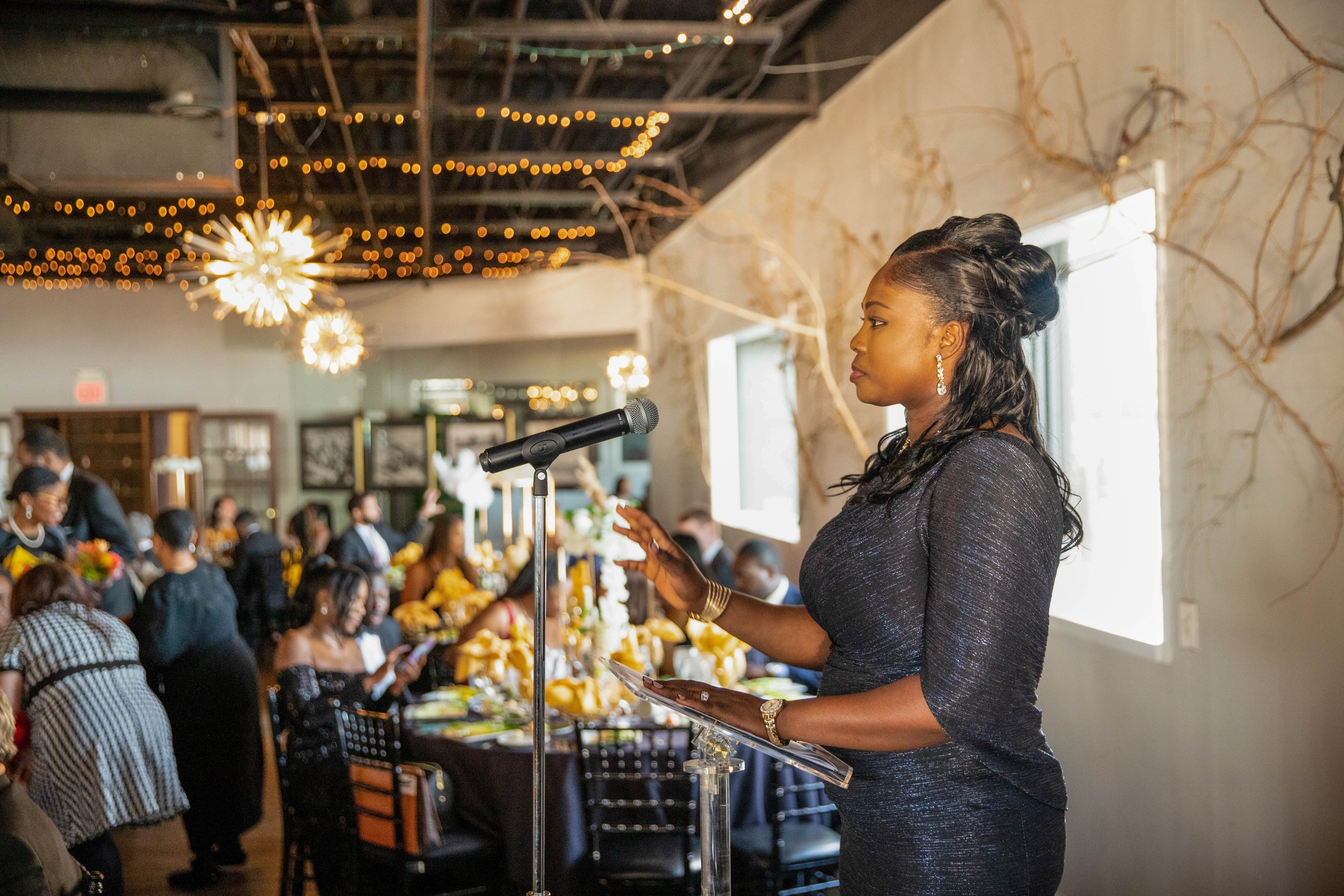 An African American woman in a dark, sparkly dress is speaking at a microphone at a formal event or banquet. She is standing in front of a large group of seated attendees, and the room is decorated with string lights and a rustic, artistic wall. The 