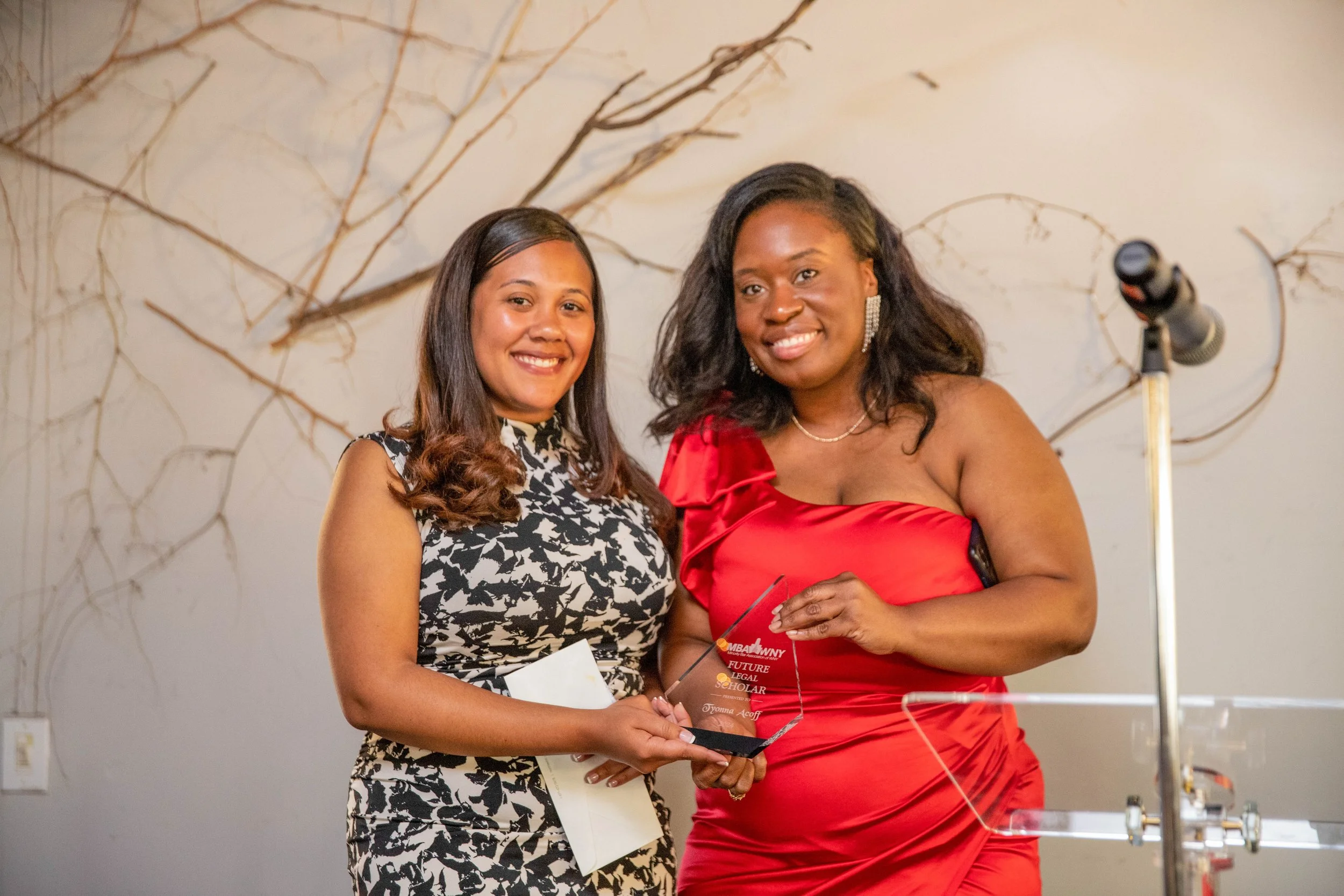 Two women at an event, one in a black and white patterned dress and the other in a red dress, holding an award or plaque together, with a microphone and a branch-decorated wall in the background.