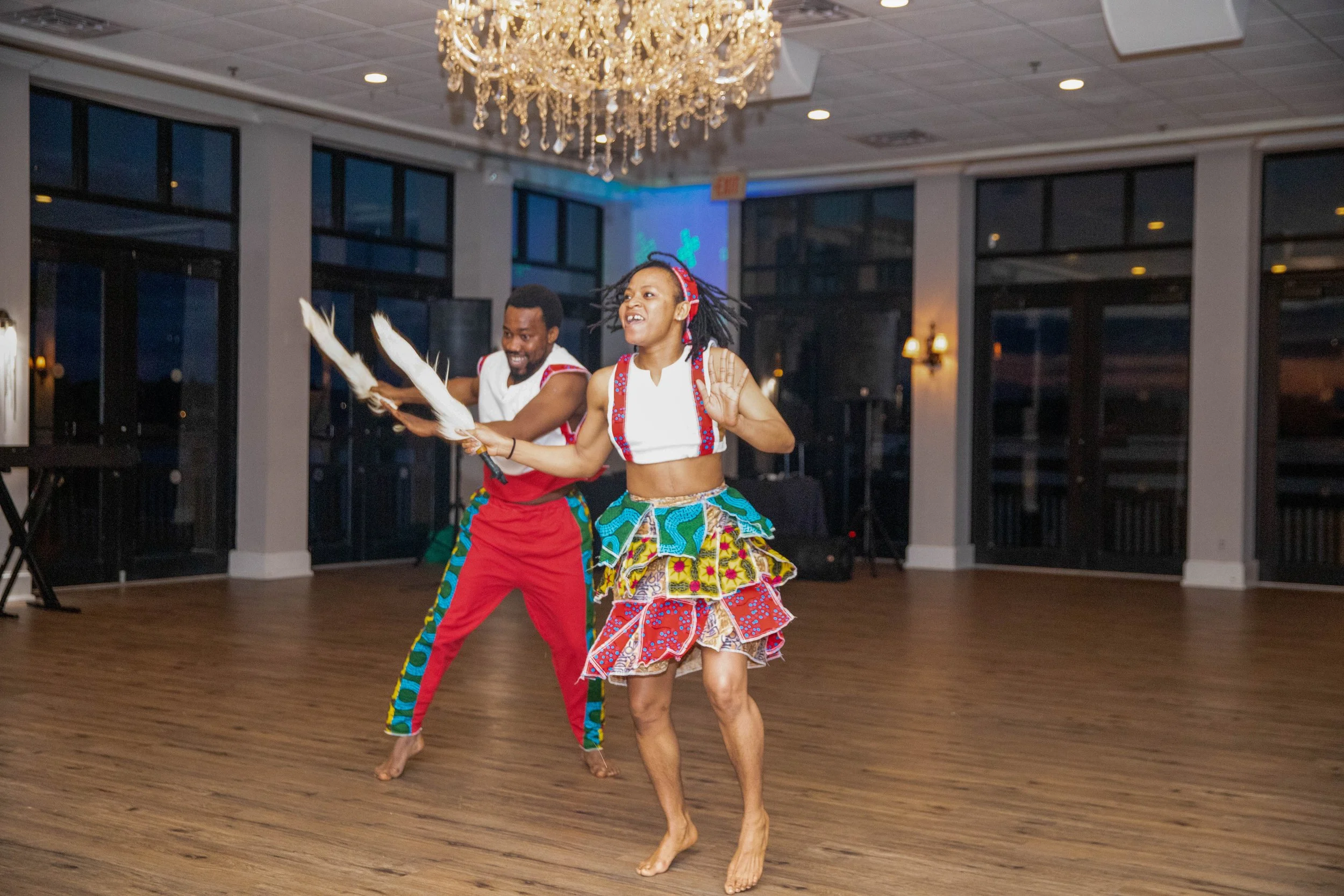 Two people dancing on a wooden floor in an elegant room with large windows, a chandelier, and a wall projection in the background. One is a woman wearing a colorful, patterned skirt and top, and the other is a man in red pants and a white shirt. They