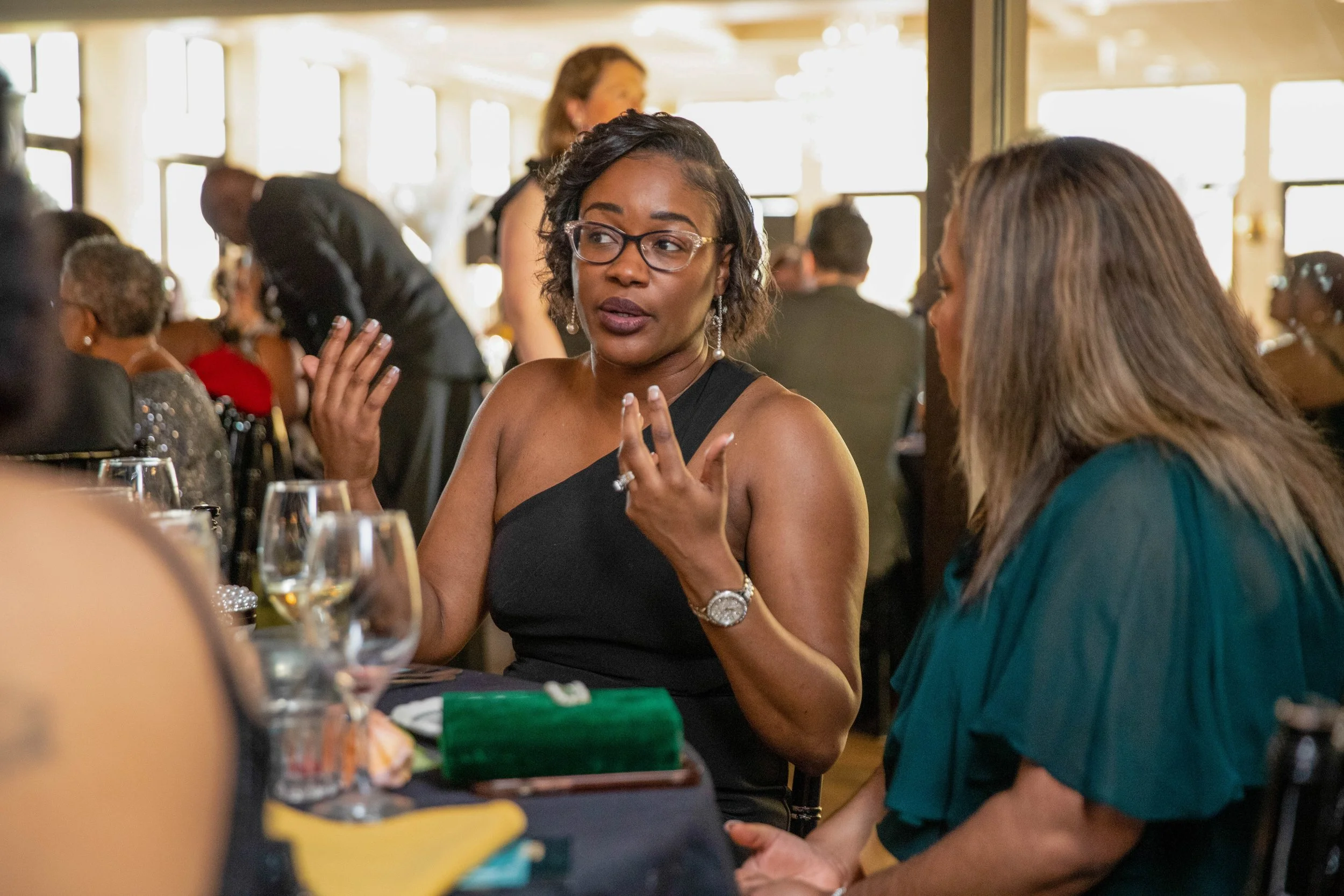 A woman wearing glasses in a black dress is talking to another woman in a teal dress at a crowded event, with tables set with drinks and glasses.