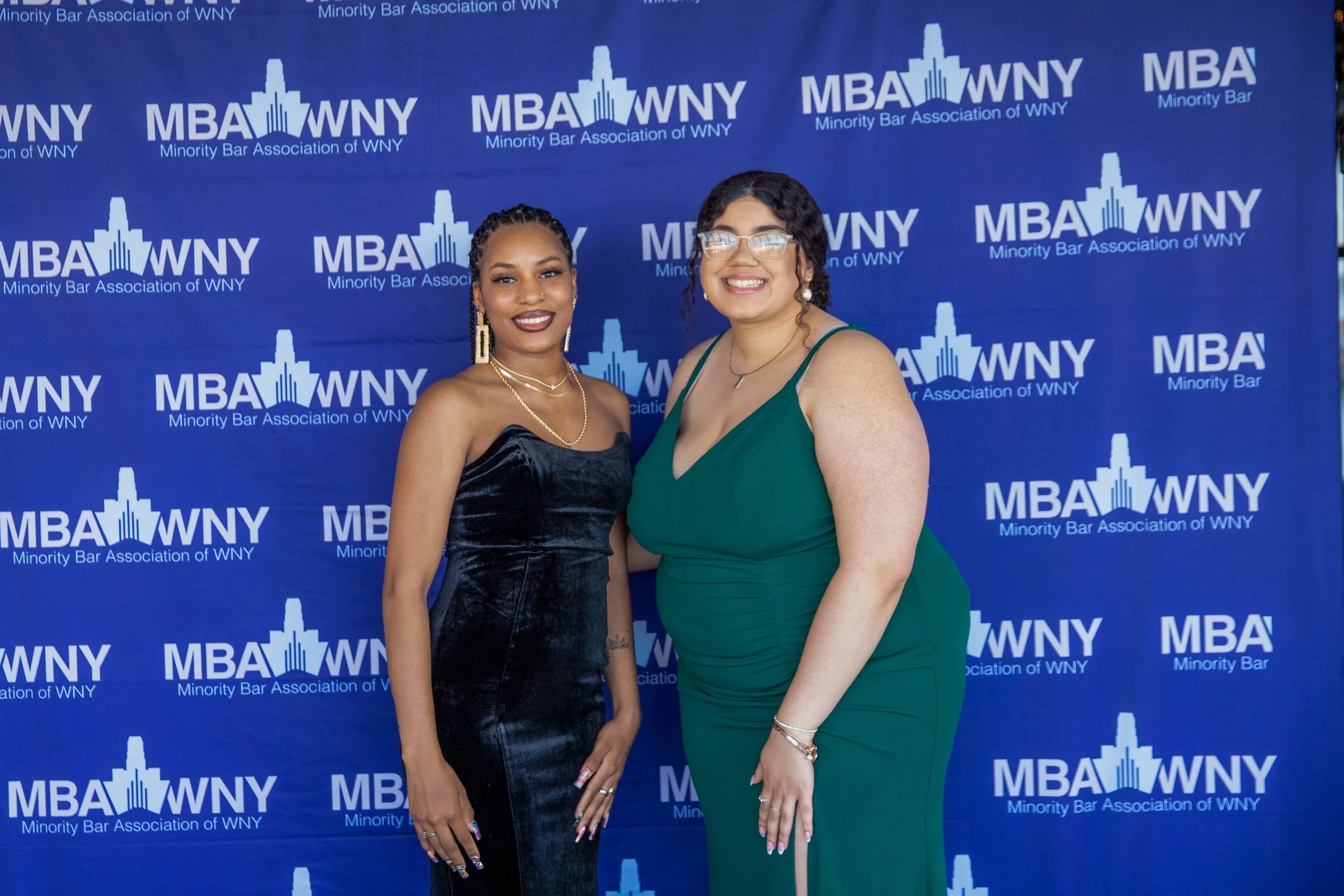 Two women standing in front of a blue backdrop with the logo and text of the Minority Bar Association of WNY, smiling at the camera. One woman is wearing a dark strapless dress with jewelry, and the other is wearing glasses and a green dress.