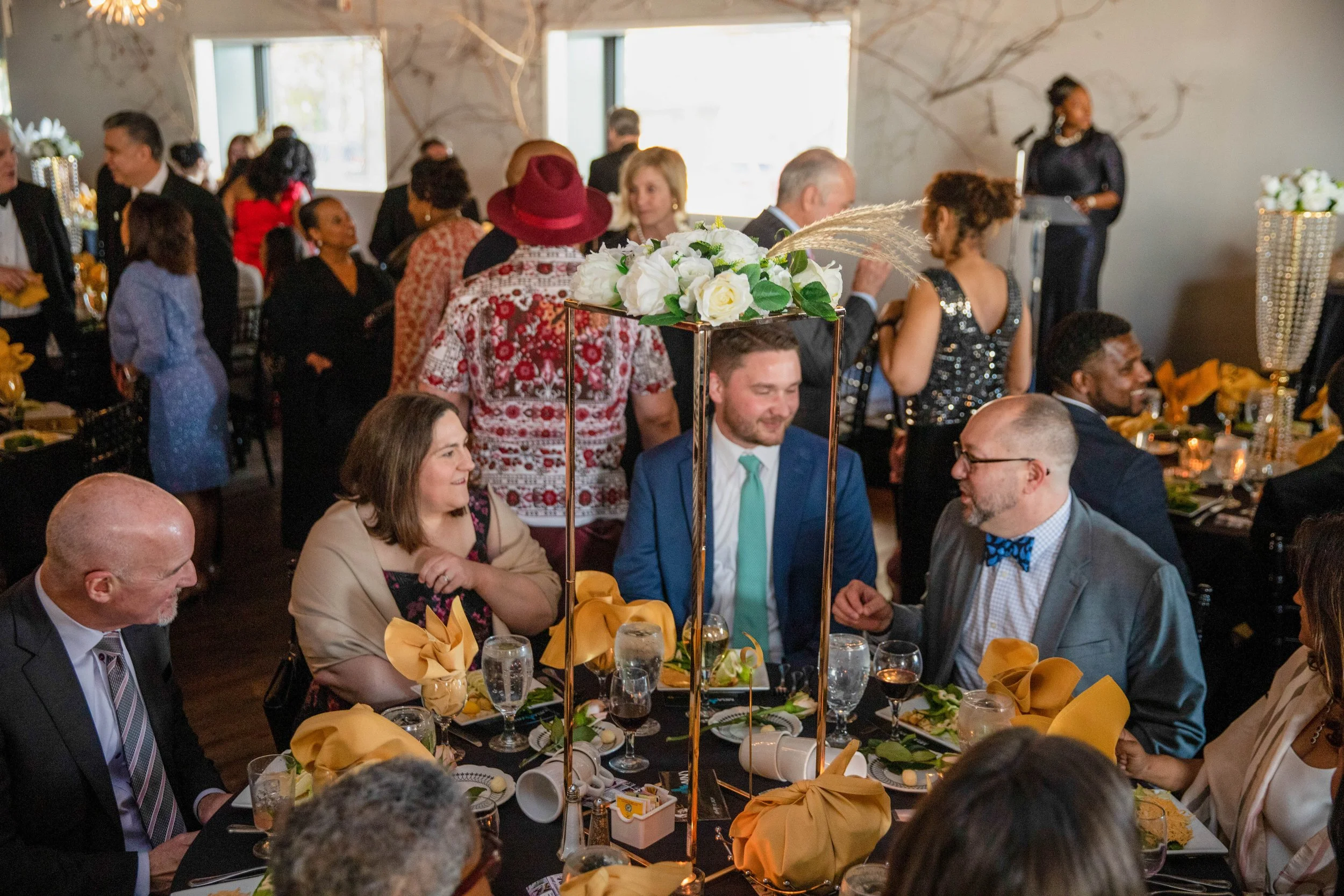Guests sitting at a long banquet table decorated with tall floral centerpieces, silverware, and yellow napkins, attending a formal event in a well-lit room with large windows and elegant wall decor. A woman in black is giving a speech at a podium.