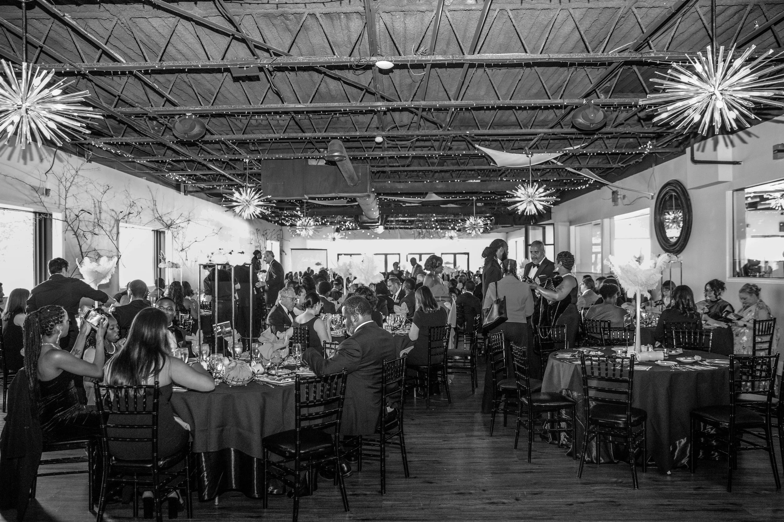A crowded indoor event or banquet with people sitting at tables, some engaged in conversations, all dressed in formal attire. The ceiling is decorated with string lights and starburst-shaped light fixtures, and the room has large windows letting in n