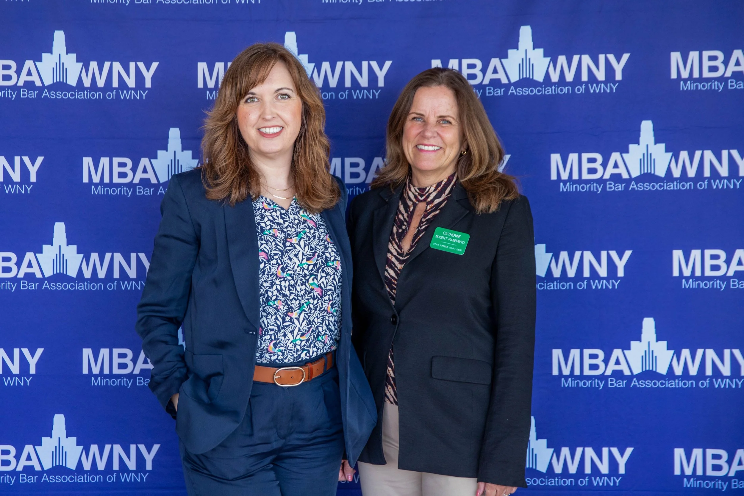 Two women standing in front of a blue backdrop with the logos of the Minority Bar Association of WNY. The woman on the left has shoulder-length curly reddish hair and is wearing a navy blue blazer over a floral shirt, with a tan belt. The woman on th
