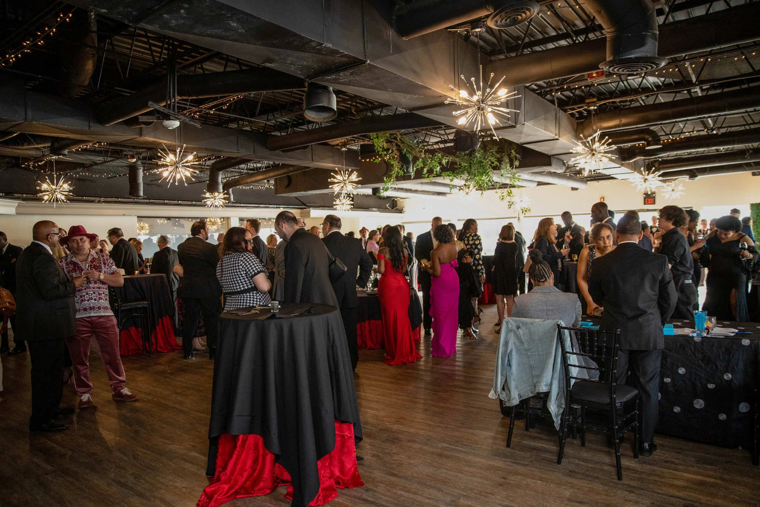 People dressed in formal attire gathering at a reception or social event inside a decorated venue with modern light fixtures and greenery.